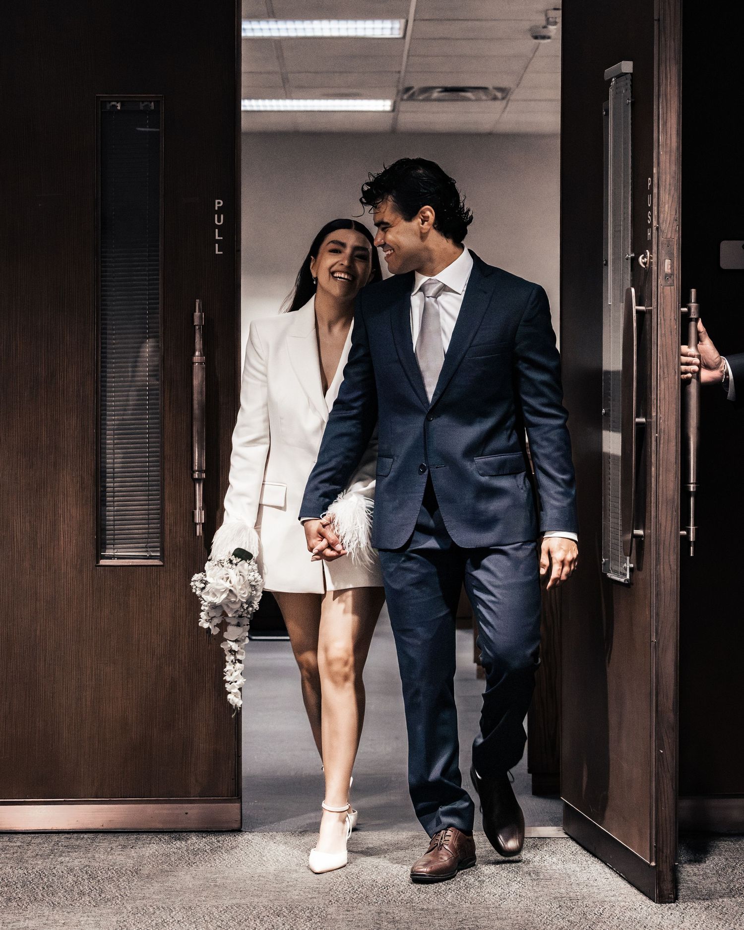 Couple in wedding attire walking hand-in-hand through a doorway, woman in white blazer dress holding bouquet, man in navy suit.