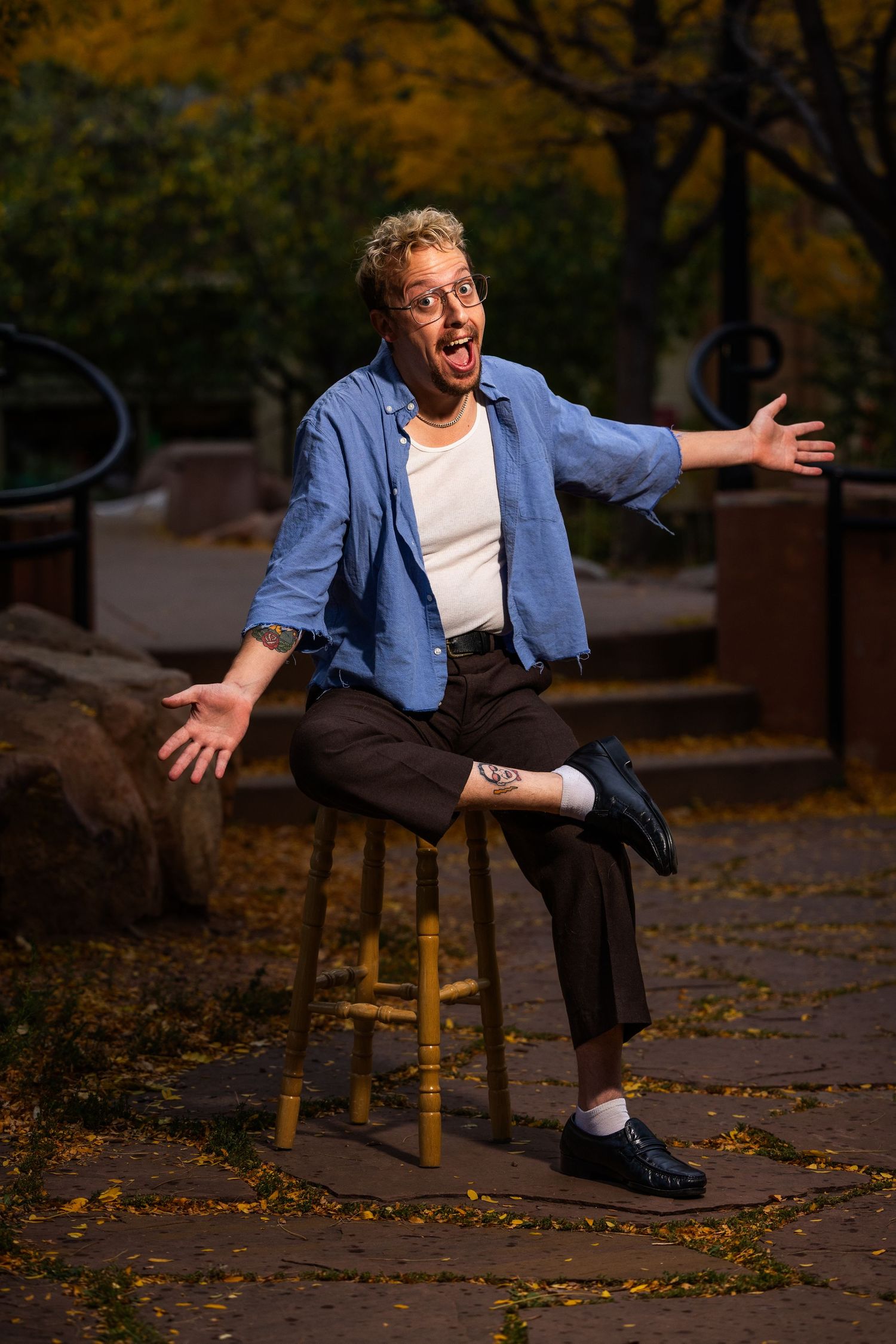 A person in a denim jacket and white shirt performs a playful falling motion sequence on a wooden stool in autumn.