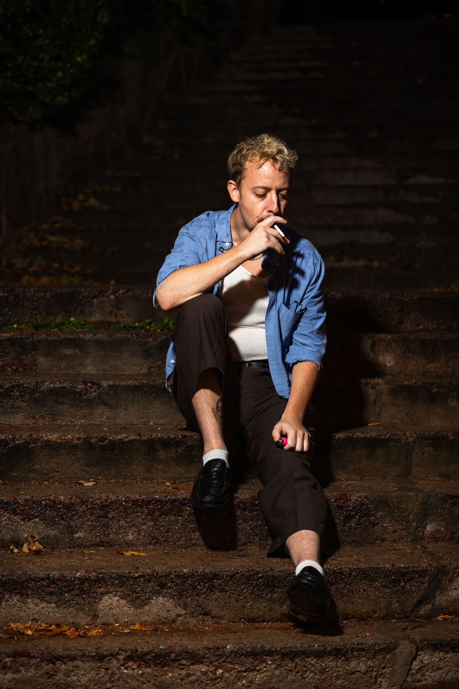 Person in denim shirt and black pants sits on outdoor stone steps in moody lighting sequence.