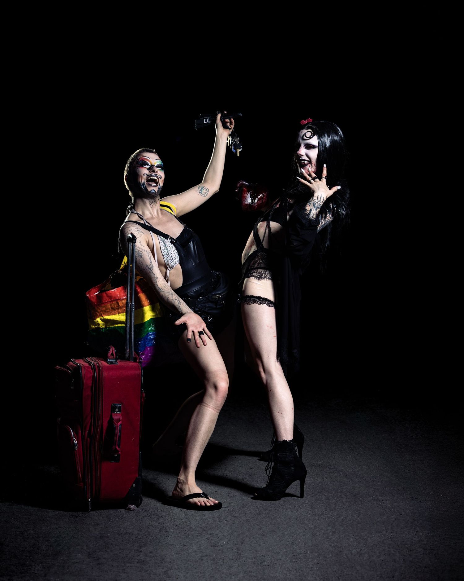 Two performers in lingerie celebrate with a rainbow flag and red suitcase against a dark background.