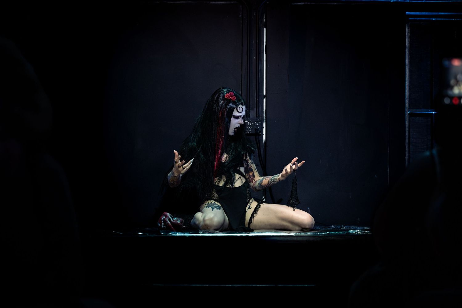 Performer in gothic costume and face paint kneels dramatically on stage under dark theatrical lighting.