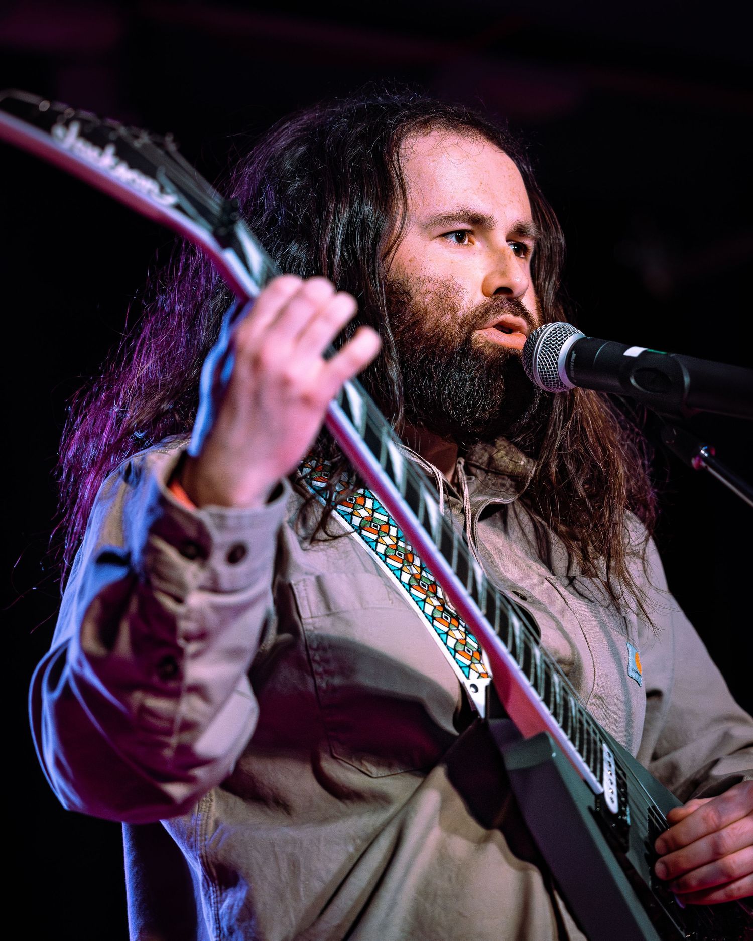 Bearded male guitarist with long hair performs on stage, playing electric guitar near a microphone.
