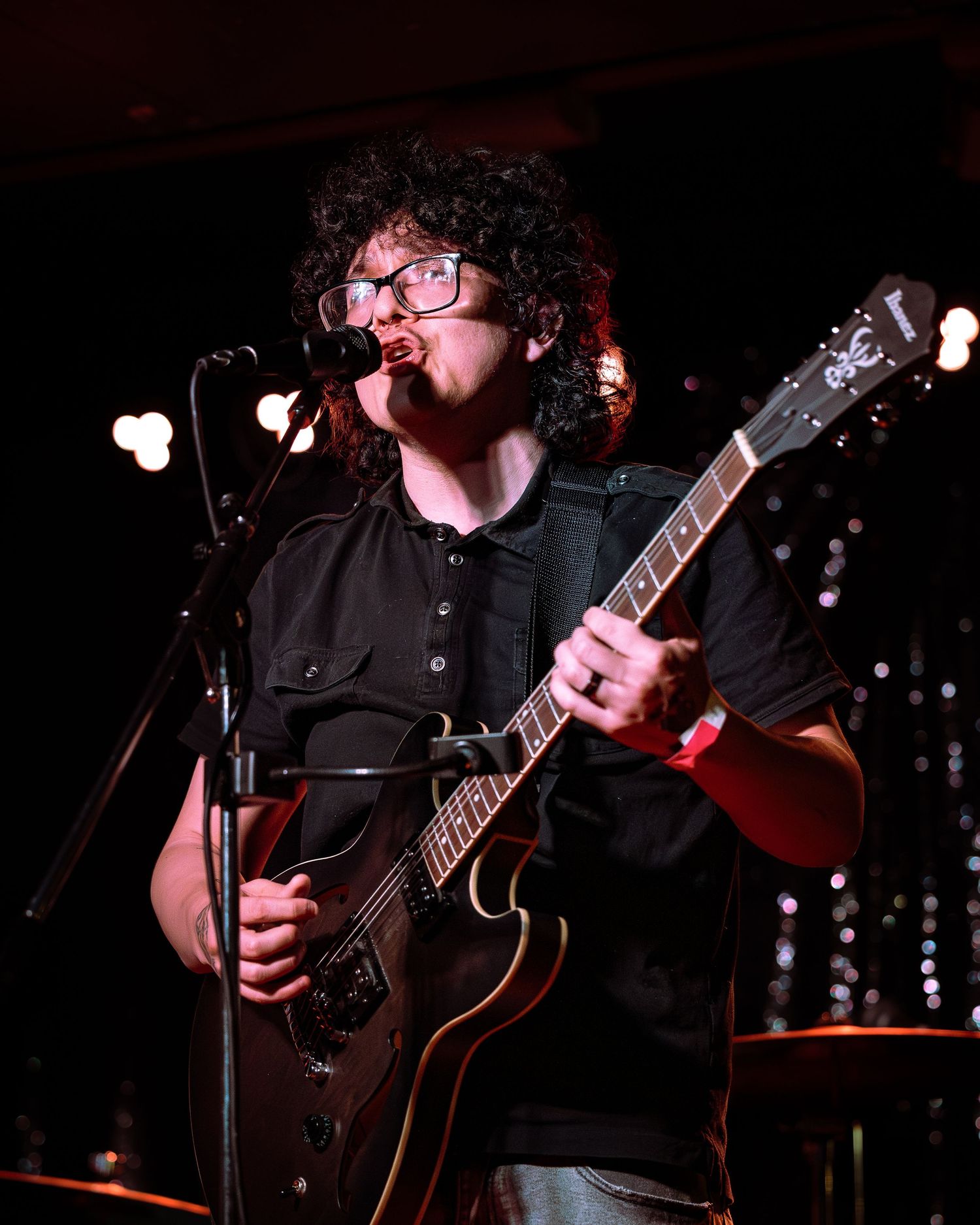 Curly-haired male guitarist with glasses performs on stage, playing a dark electric guitar at a live music venue.