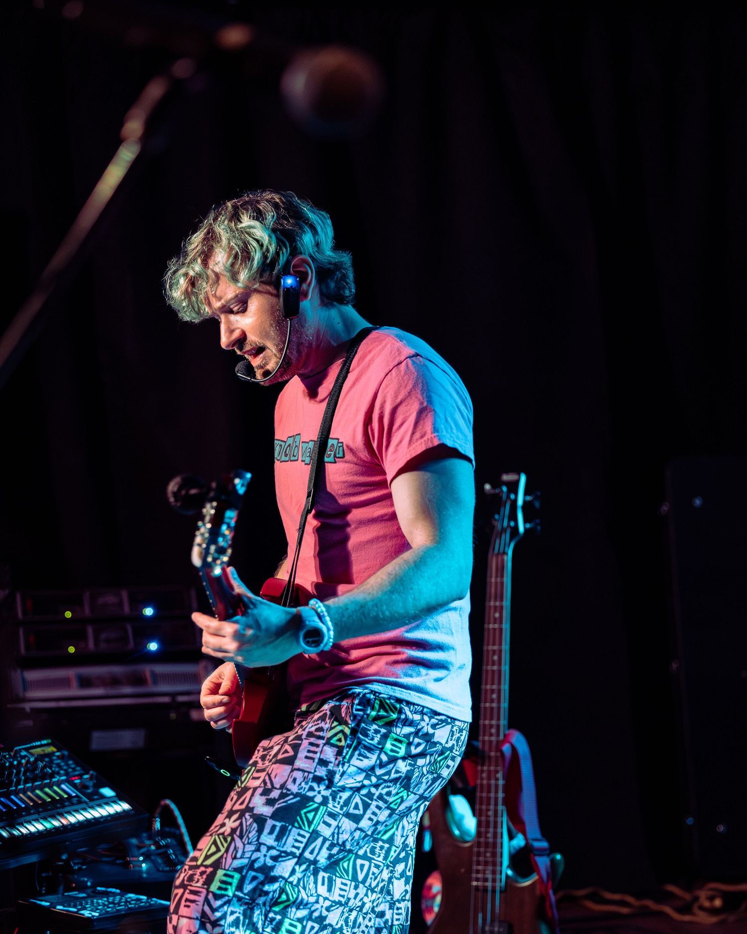 Guitarist in pink shirt and patterned pants plays electric guitar on stage under colorful lights.