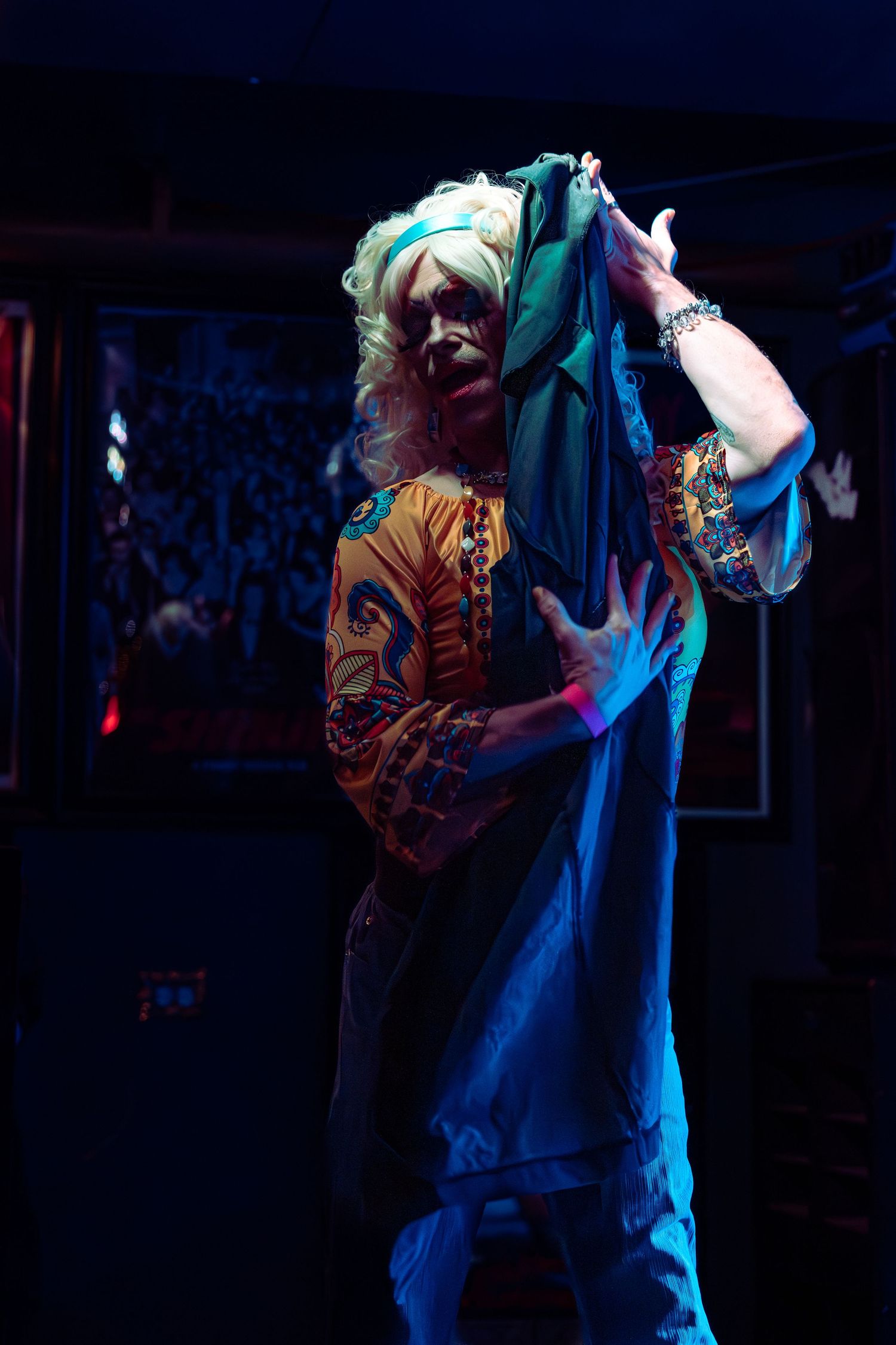 Drag performer in white wig holds teal fabric overhead on a dark stage with blue lighting.