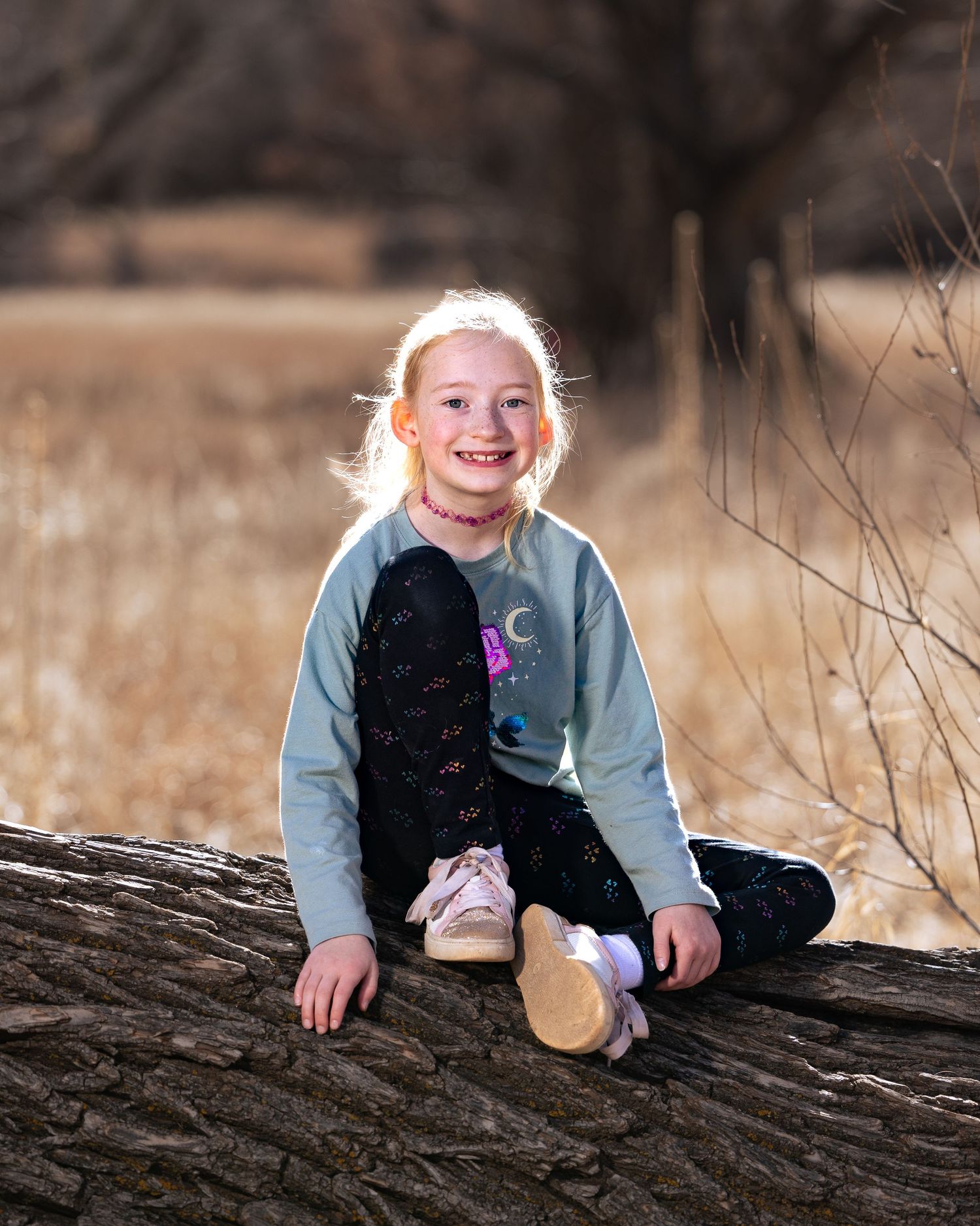 A smiling blonde girl sits on a log in a sunlit field, wearing a mint green top and black leggings.