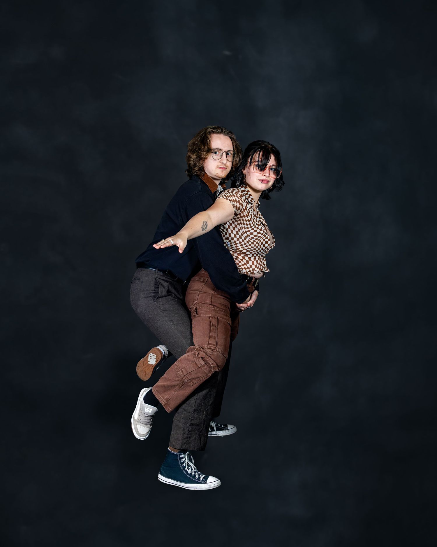 Two young people pose playfully in a studio, one giving the other a piggyback ride against a dark background.