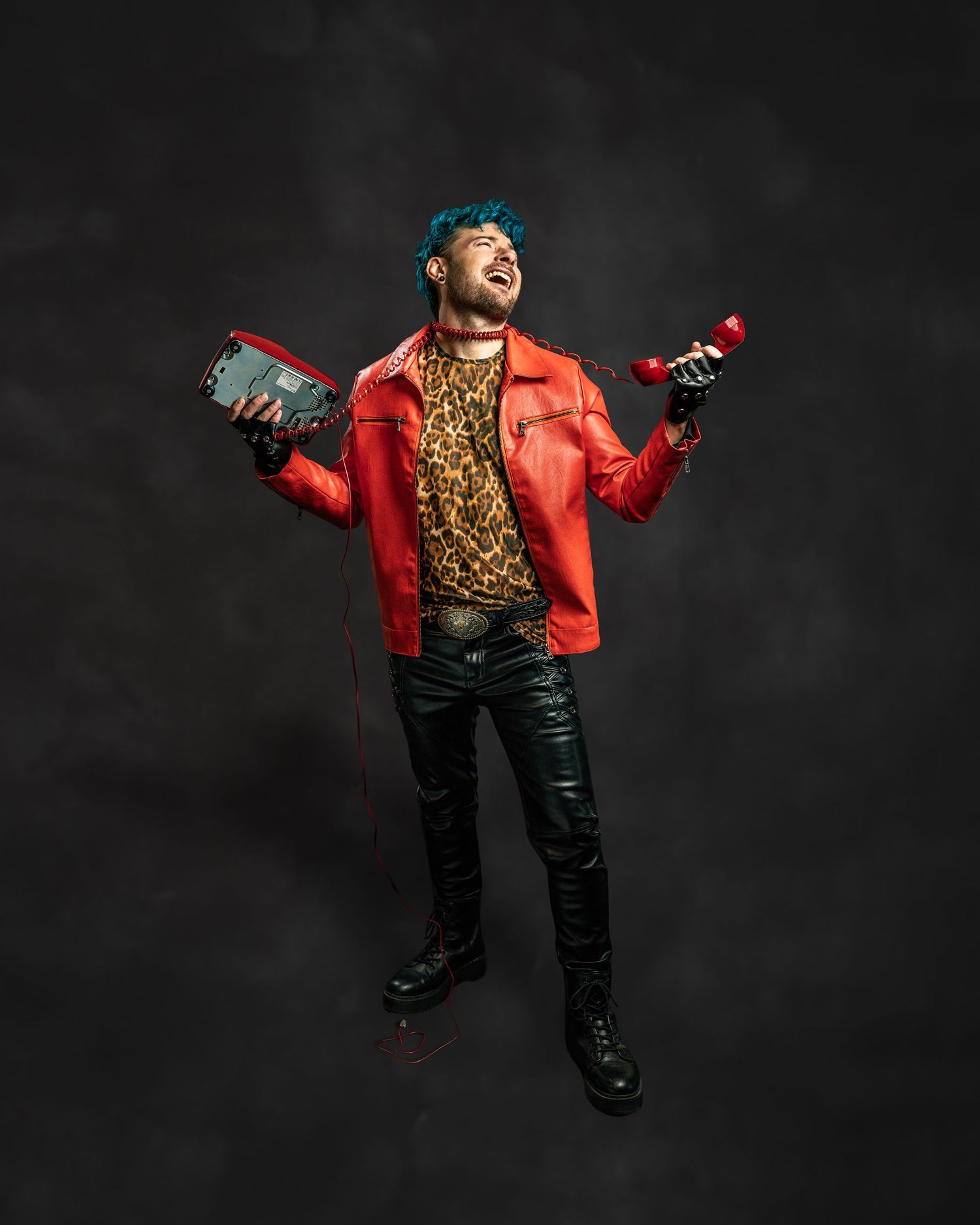 Man with teal hair in red leather jacket holds vintage red phone and boombox, wearing leopard print shirt and black leather pants.