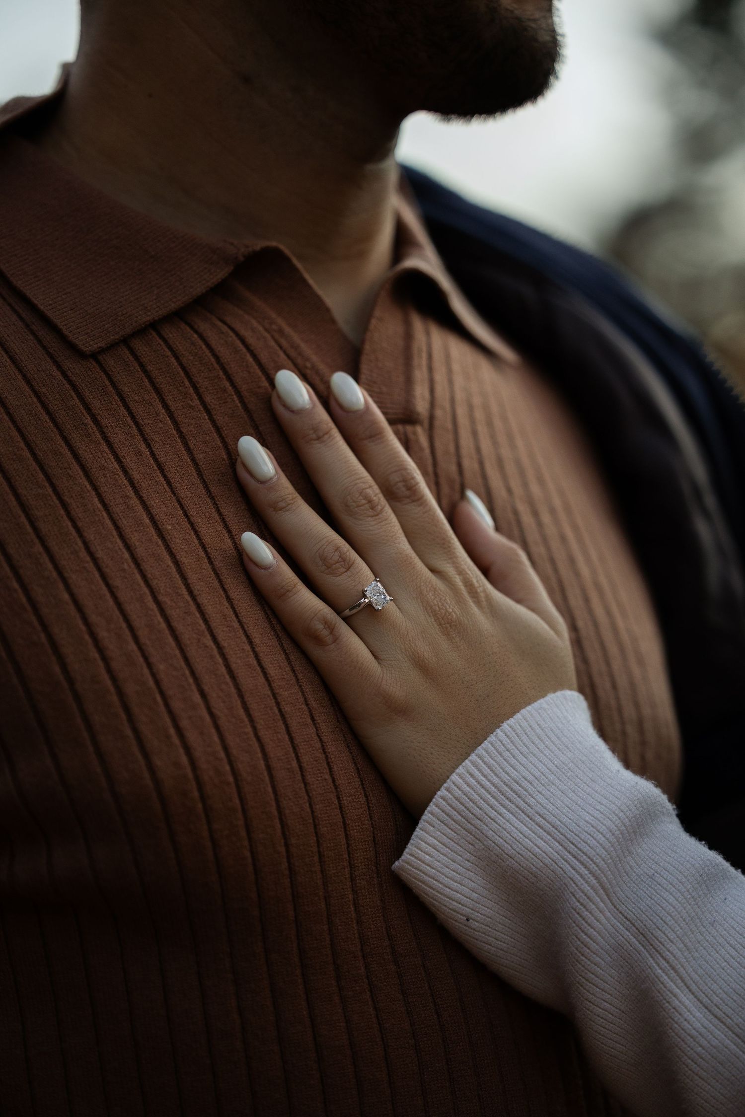 Close-up of a hand wearing an engagement ring against a brown ribbed sweater background.