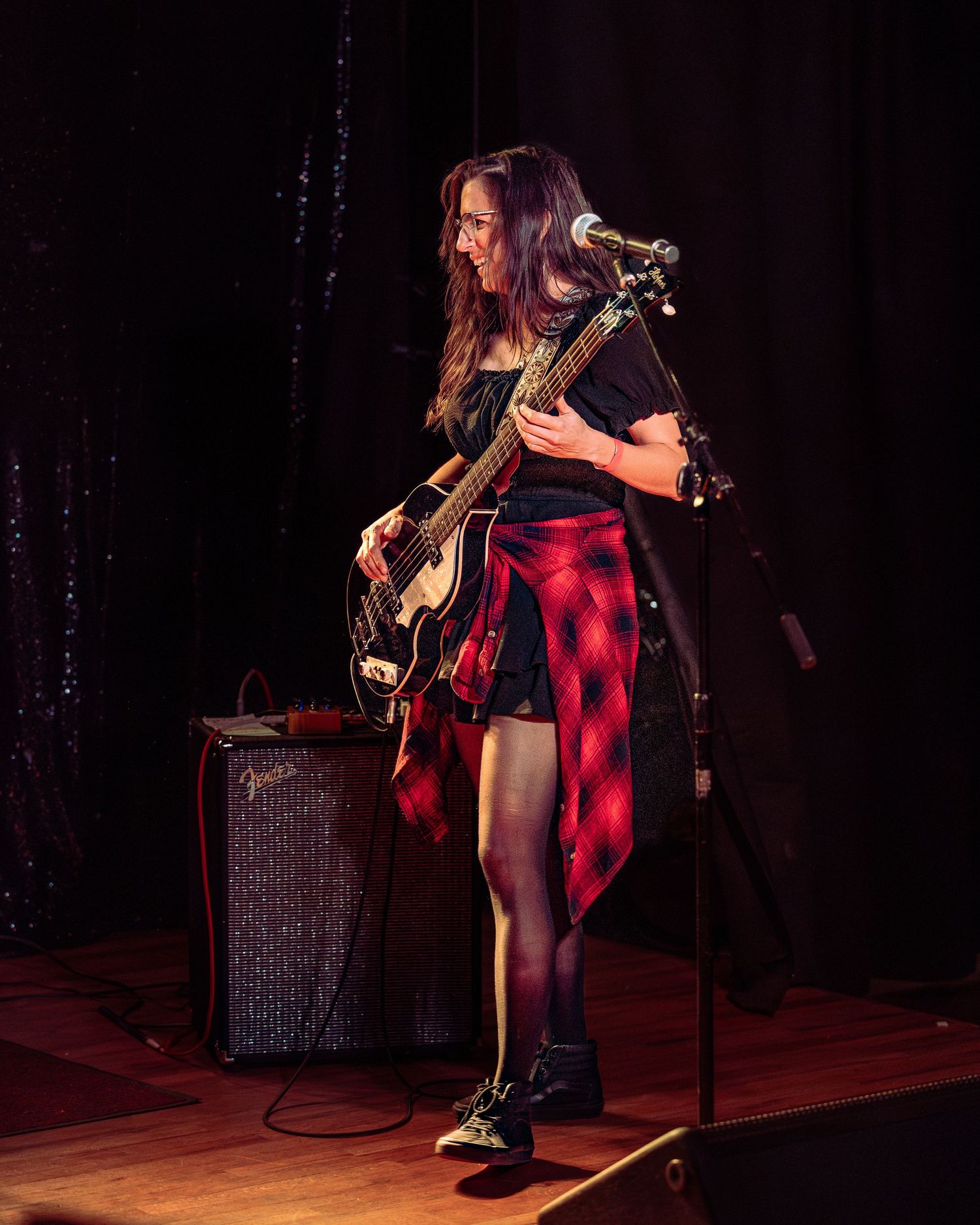 Female guitarist performing on stage, wearing plaid shirt and black outfit, playing electric guitar at live music event.