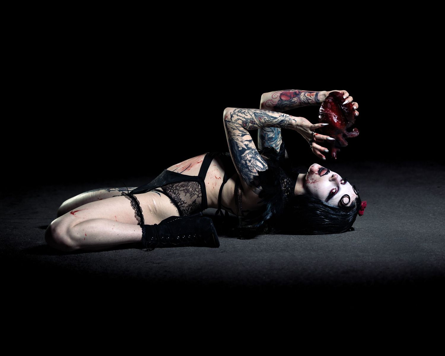 Tattooed woman in black lingerie poses dramatically on dark floor with gothic makeup and red accessories.