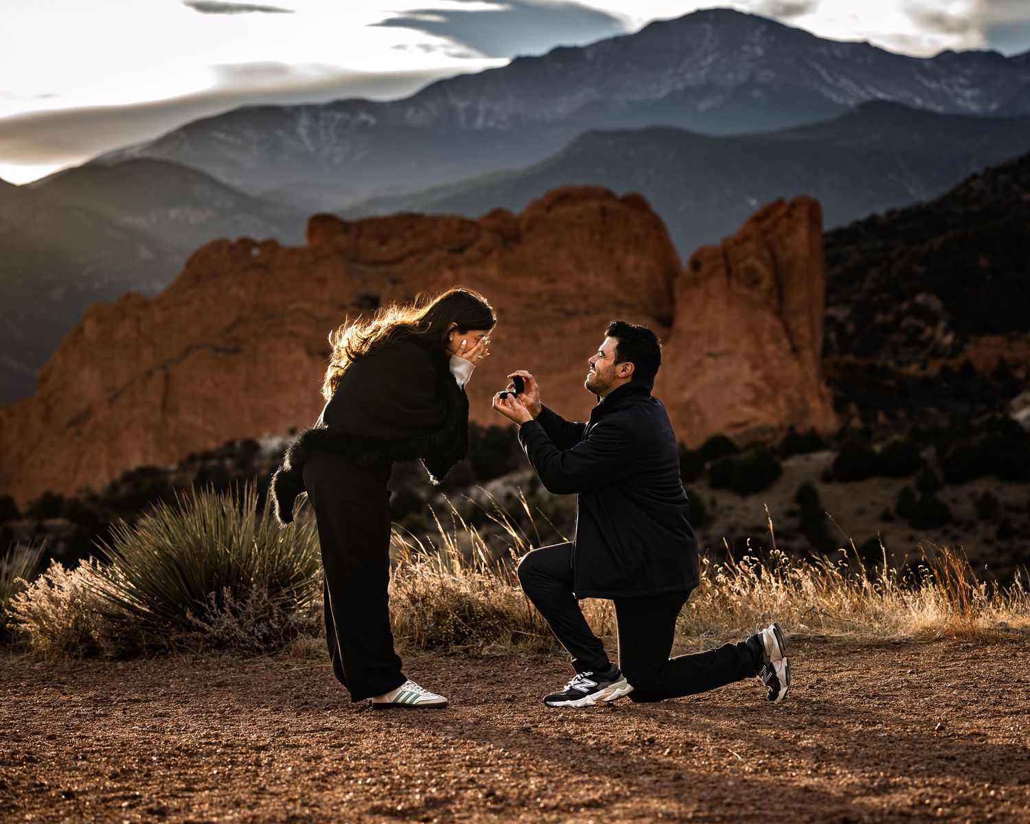 Man proposes to woman on one knee at Garden of the Gods with Rocky Mountains backdrop at sunset.
