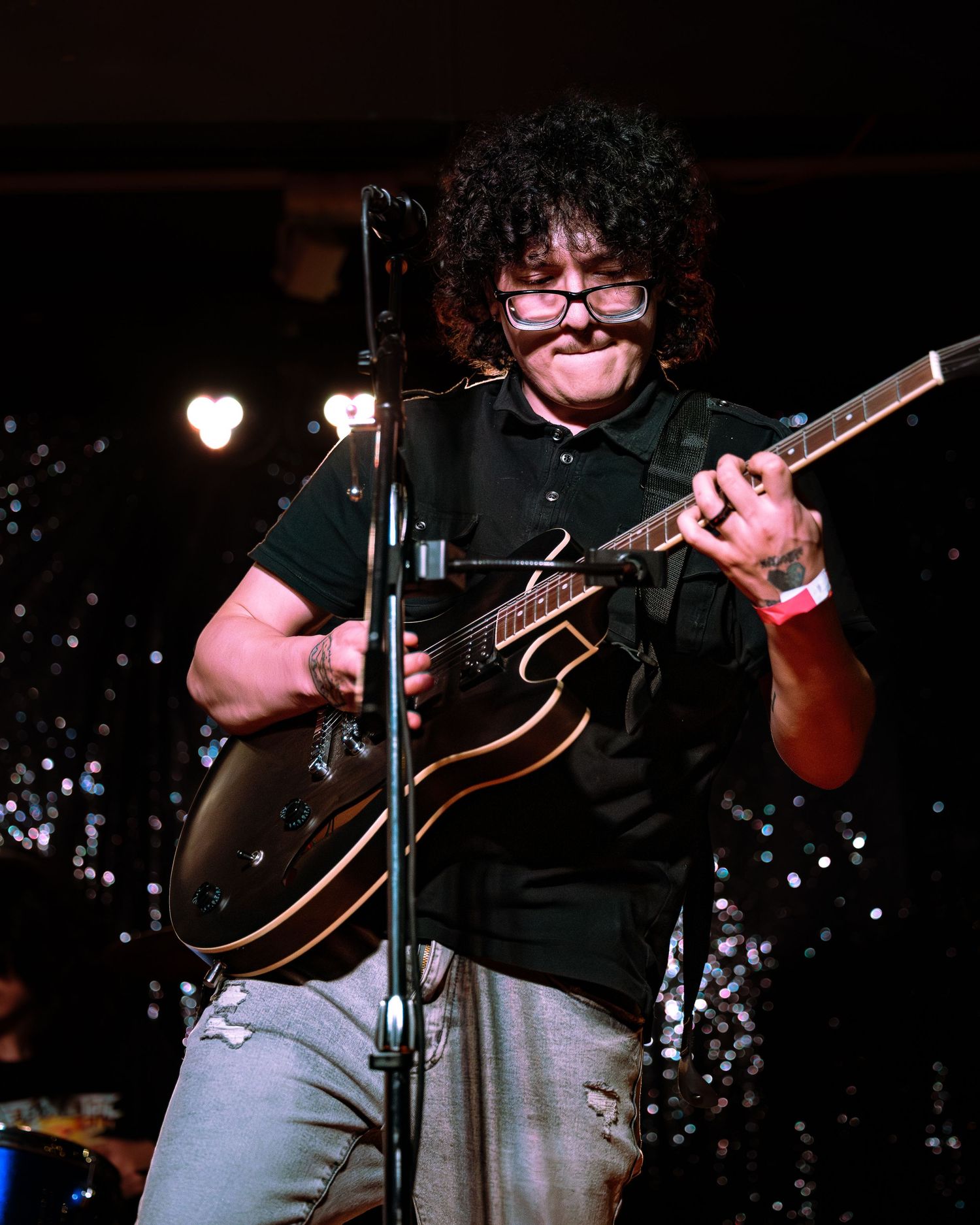 Curly-haired guitarist with glasses playing a dark hollow-body electric guitar on stage.