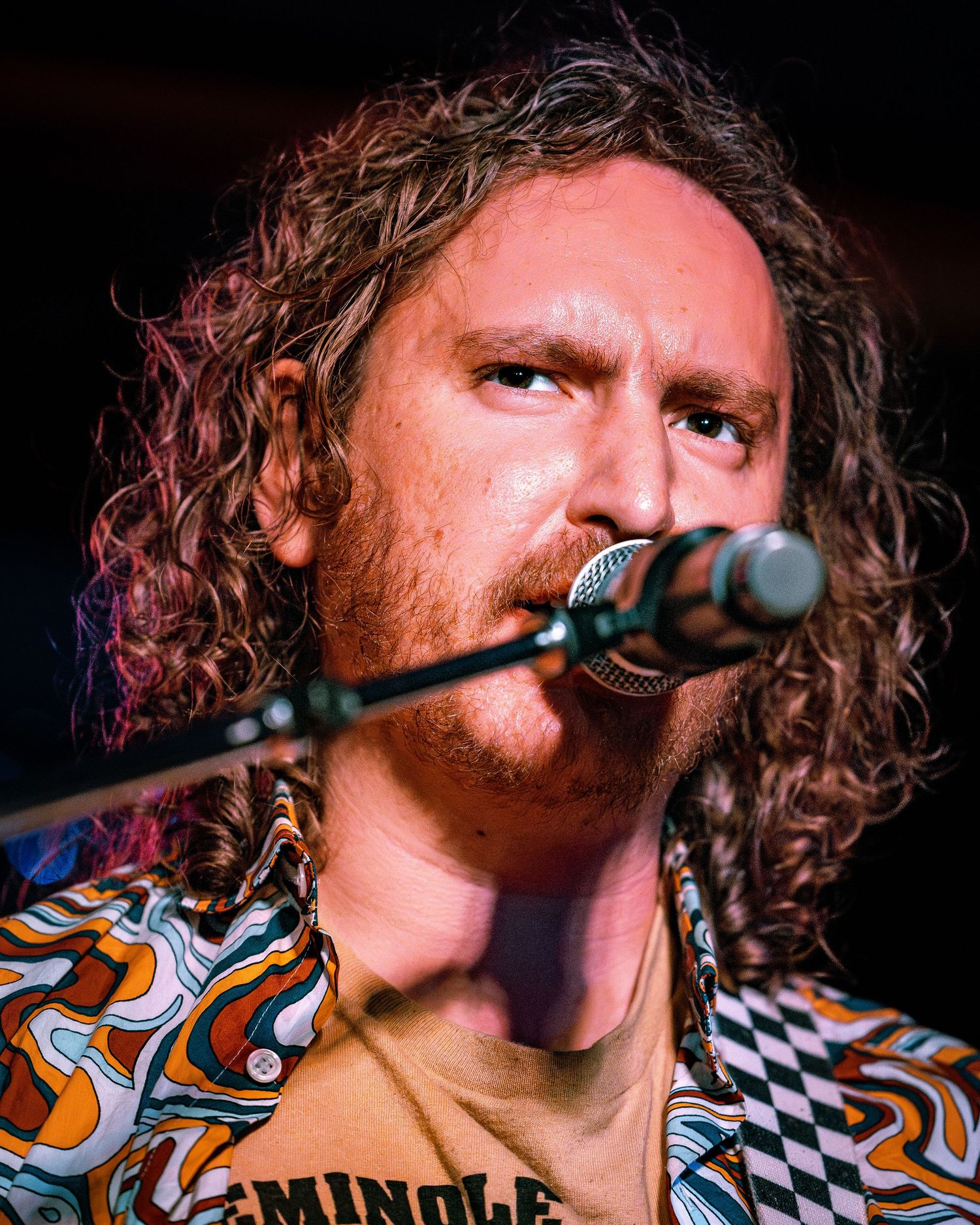 Curly-haired male vocalist sings into microphone wearing colorful patterned shirt on stage.