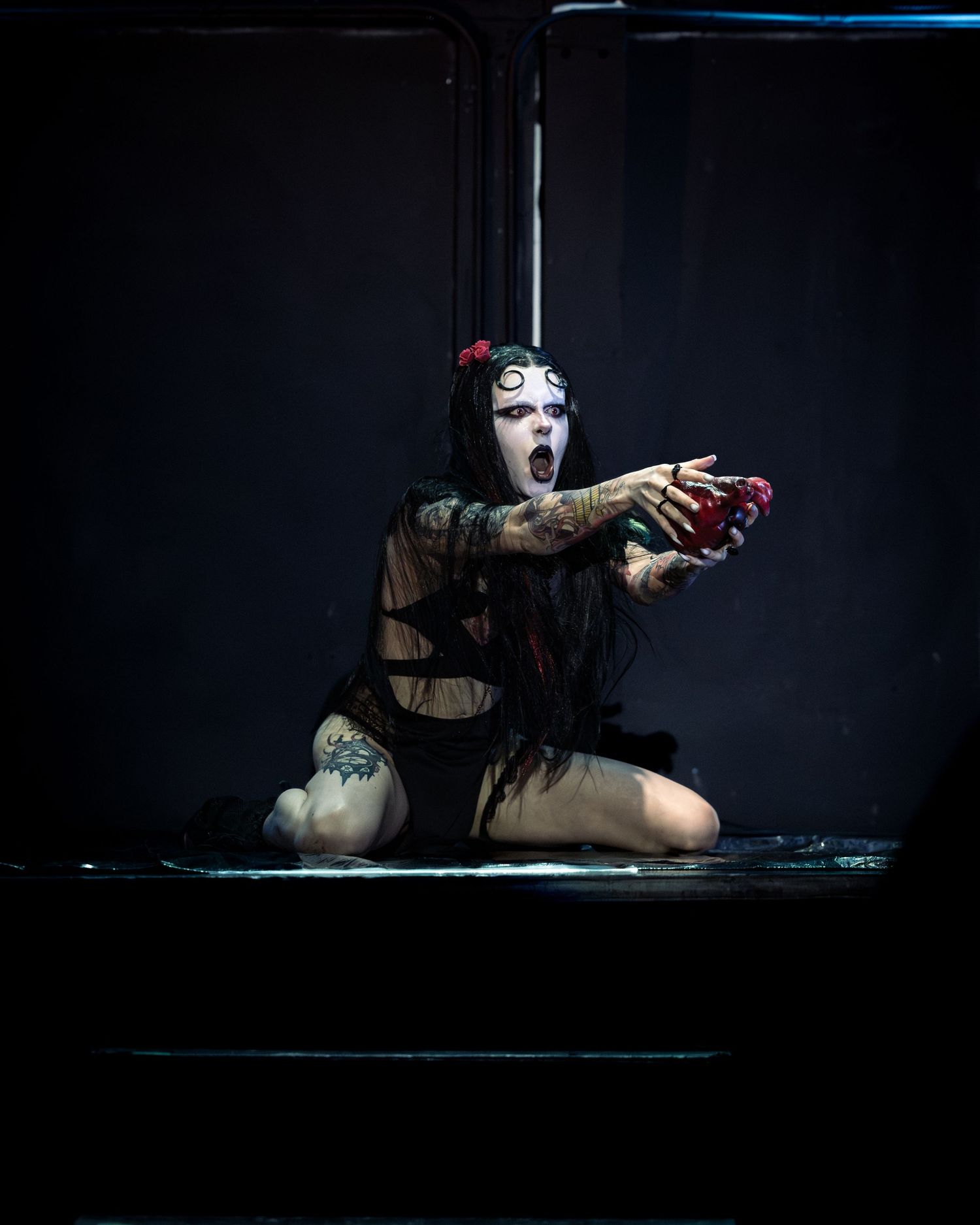 Dramatic performer in gothic makeup and dark costume kneels on stage holding a red object with mouth open.