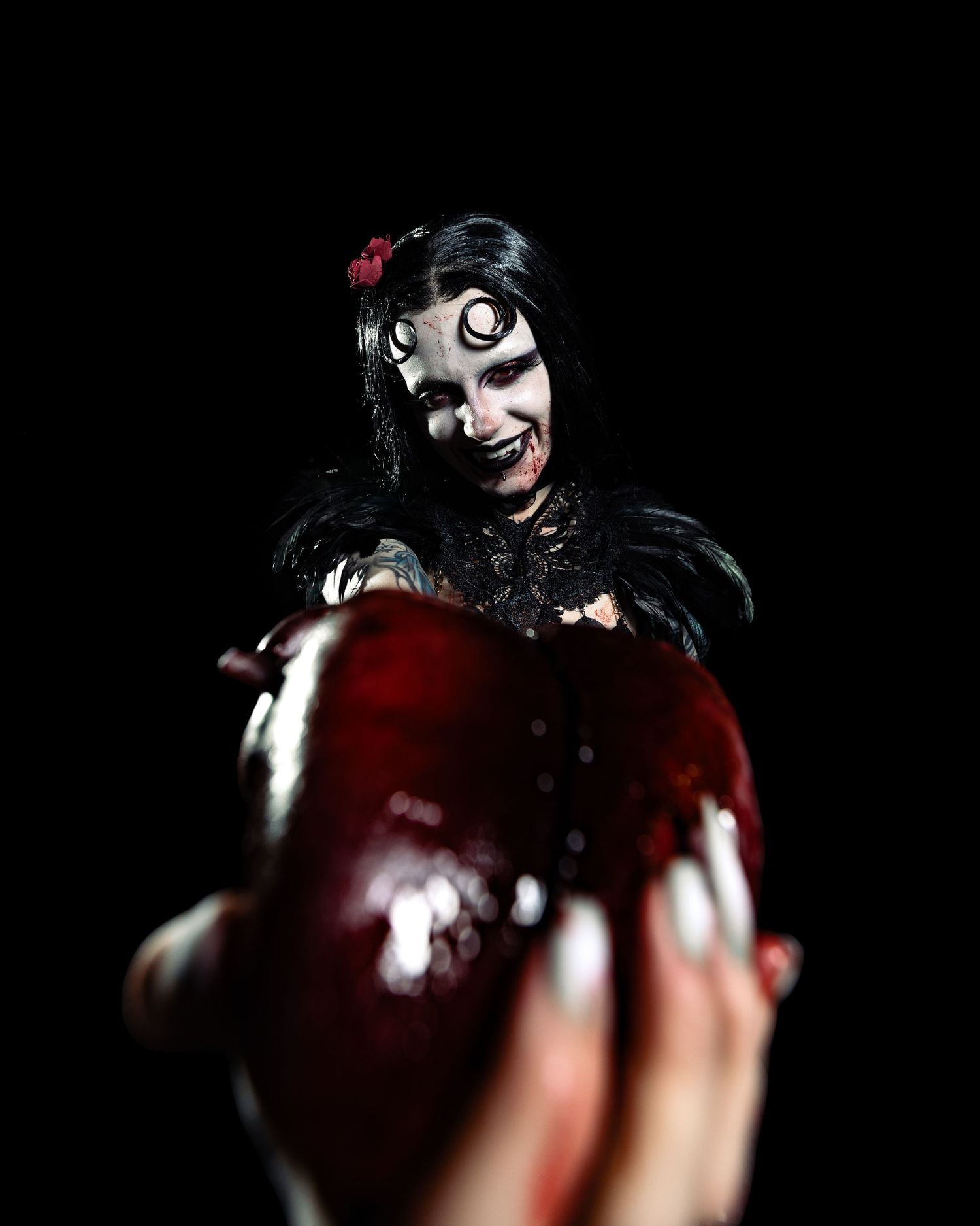 Gothic zombie-style performer with black feathers and dramatic makeup offering bloody red heart to camera.