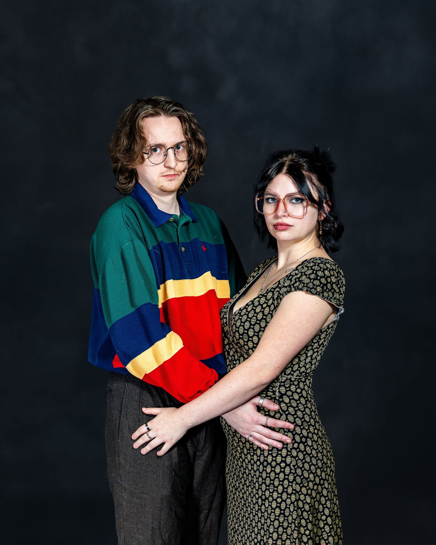 Two young adults posing together, man in colorblock sweater and woman in floral dress with glasses, studio portrait.