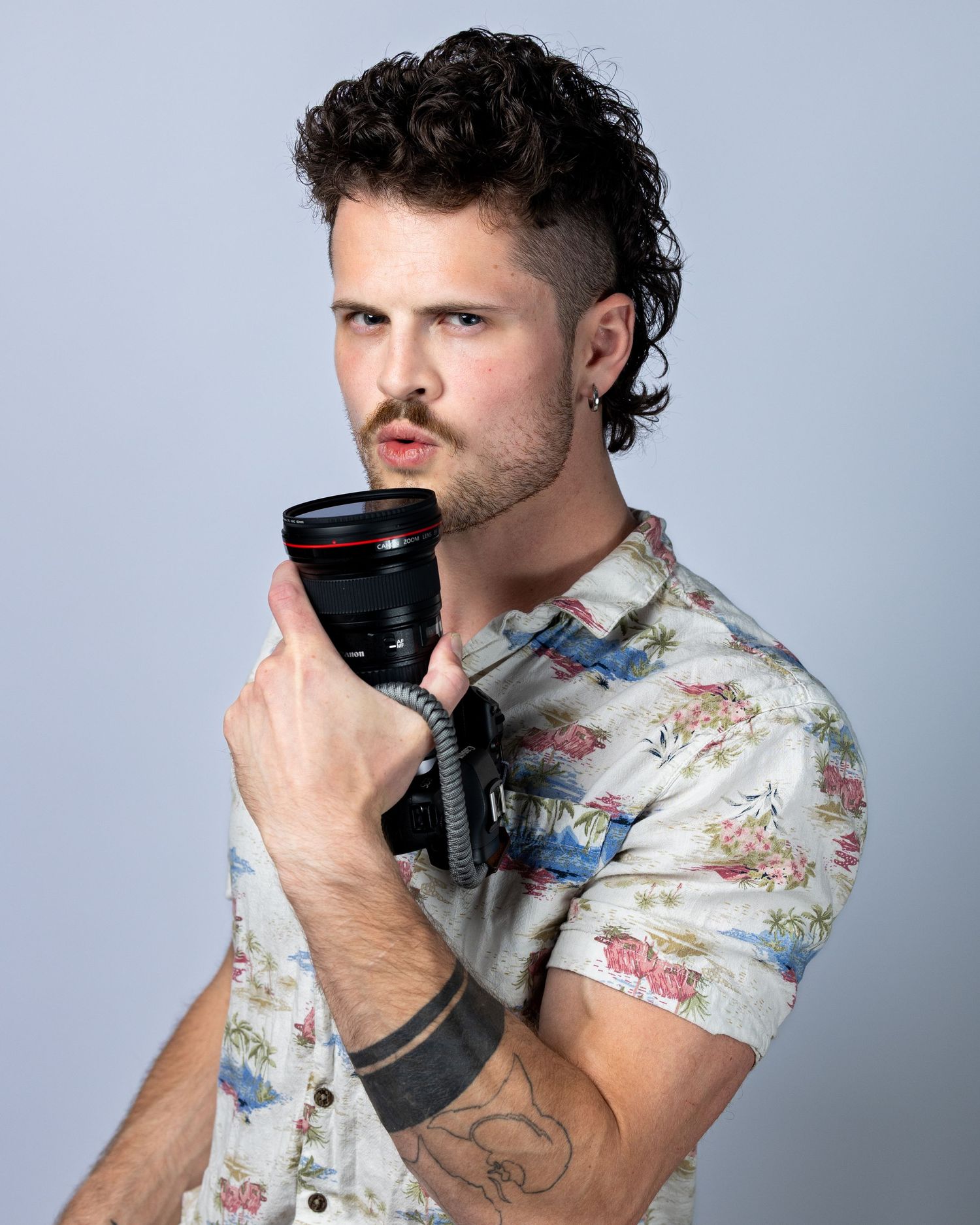 Man in floral shirt holding a camera lens close to his face, with tattoos and curly hair, studio portrait.