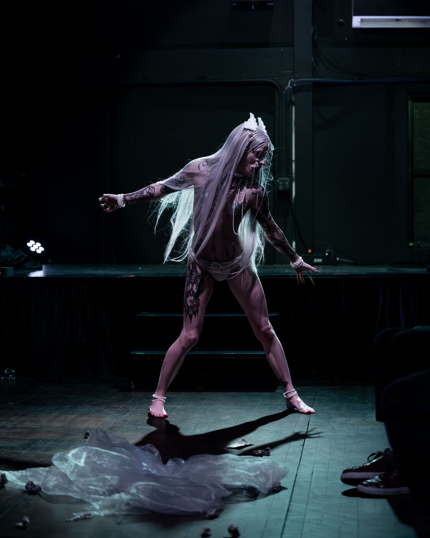 Tattooed performer with long white hair dances dramatically on a dark stage in minimal costume.