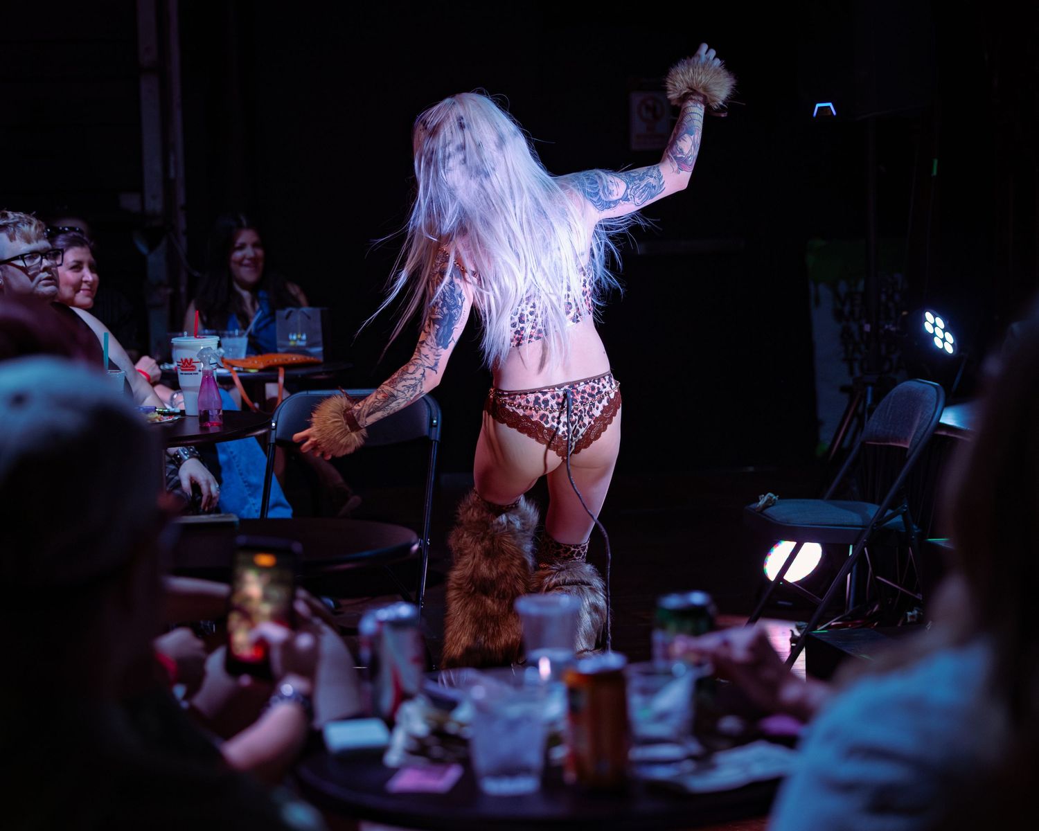Tattooed blonde performer in leopard bikini walking among audience tables during burlesque show.