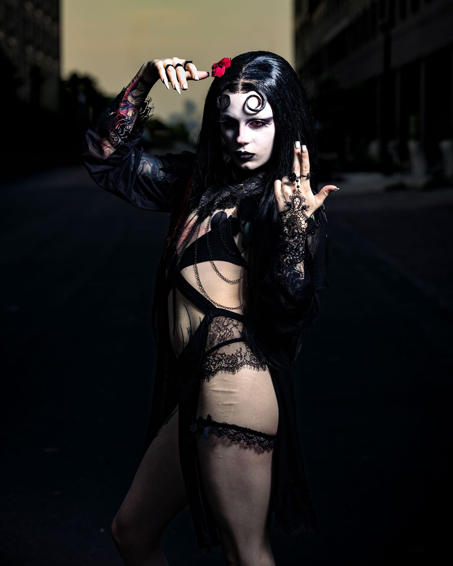 Gothic model in black lace lingerie with moon face paint poses on urban street at dusk.