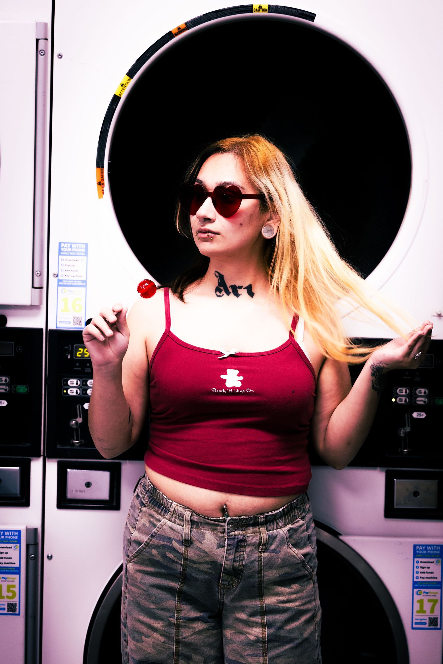 Woman in red tank top and camo pants poses confidently in front of industrial washing machines.