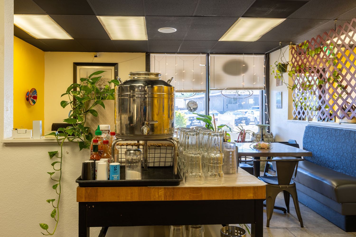 Cafe interior with a wooden beverage station, glass dispensers, plants, and bright windows with seating area.