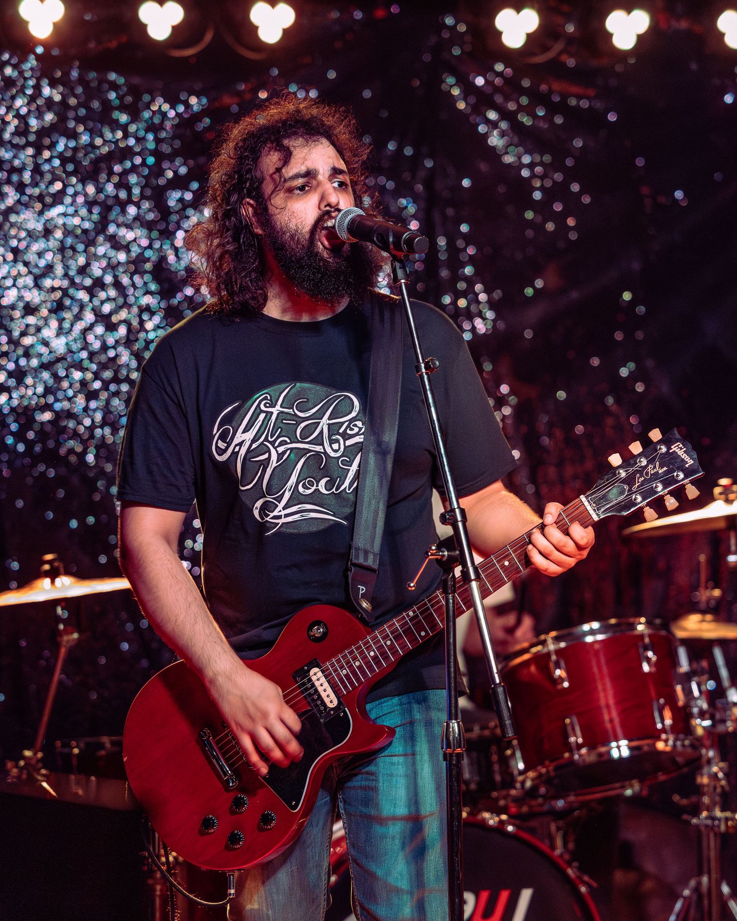 Bearded male guitarist in black t-shirt singing into microphone while playing red Les Paul guitar on stage.