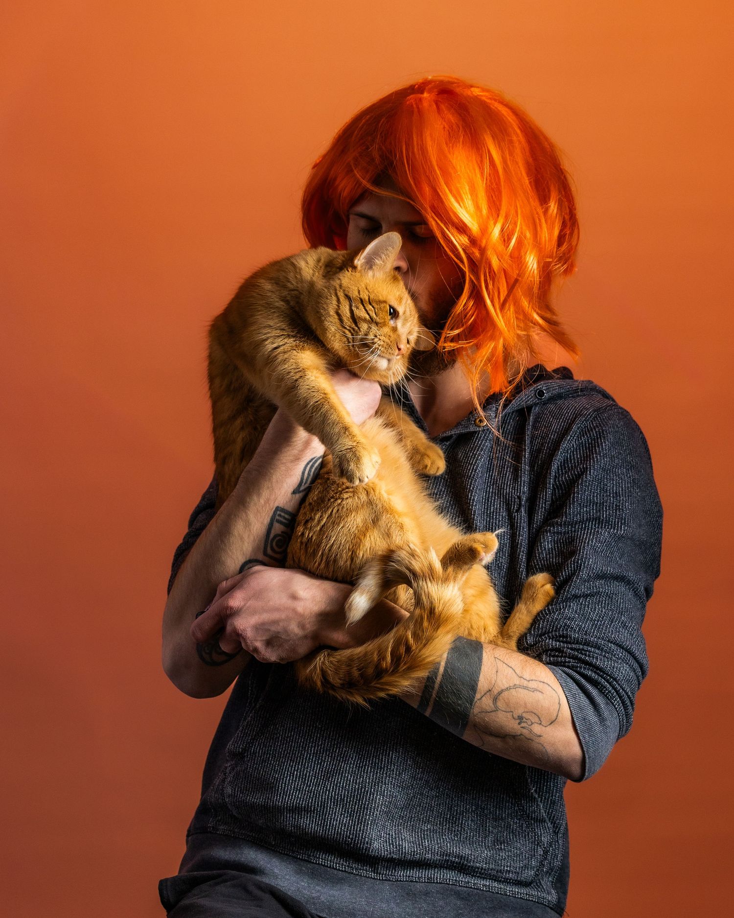 A ginger cat stretches out while being held against an orange background by someone wearing denim.