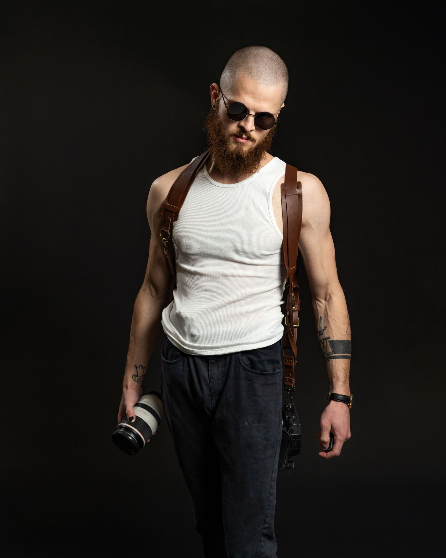 Athletic figure in white tank top and dark pants holding camera equipment against dark background with leather strap.