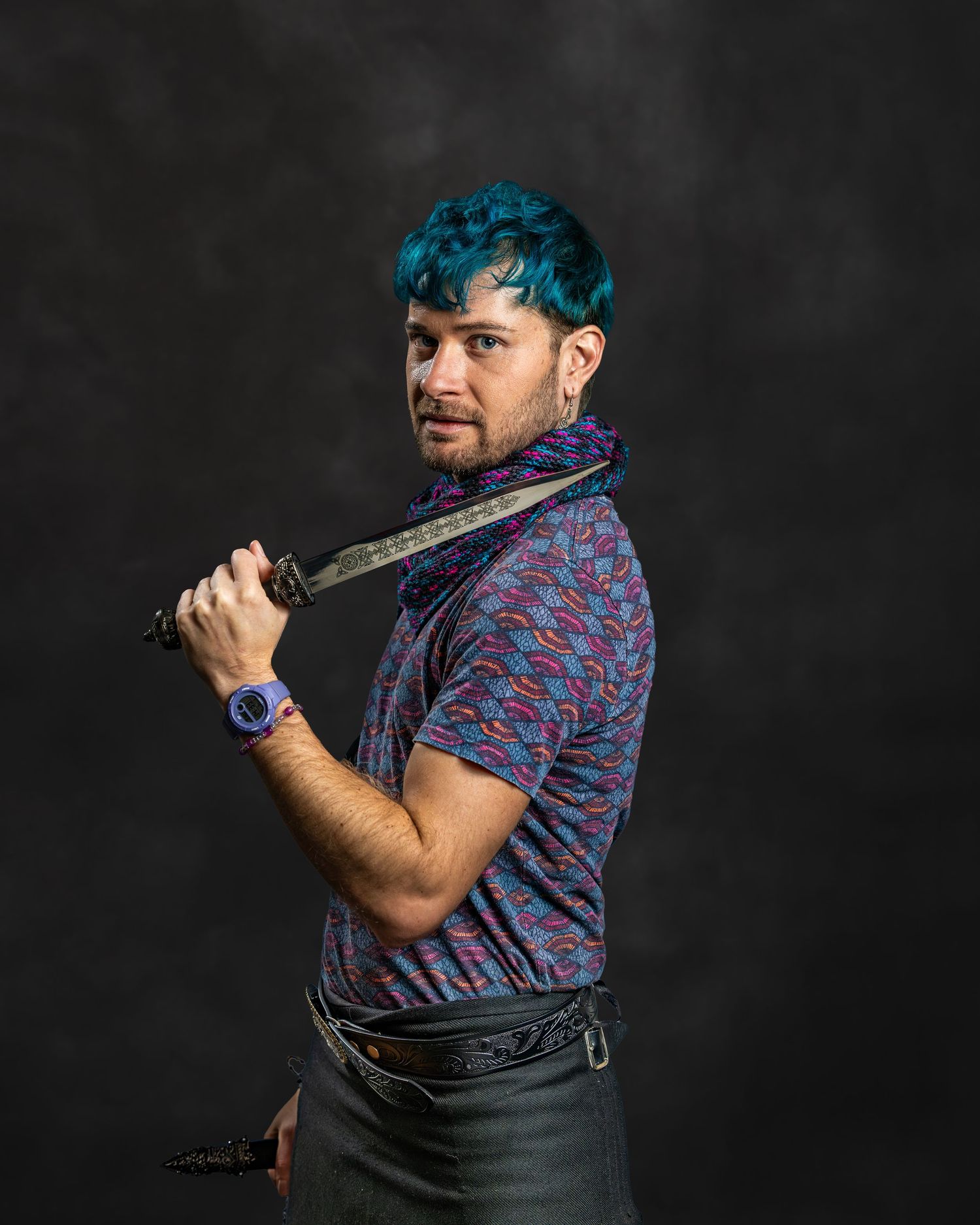 Man with teal hair holding a sword behind his back wearing a patterned shirt and leather belt.