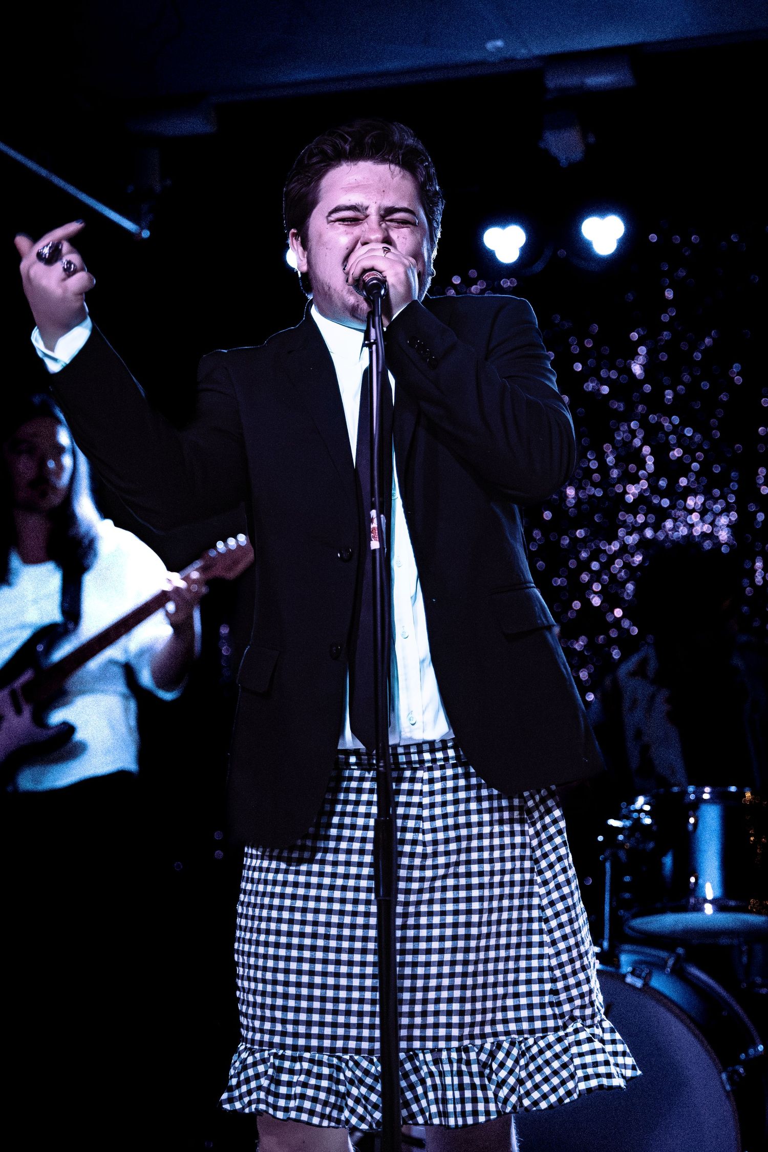 Male vocalist in a black blazer and checkered skirt singing into a microphone on stage.