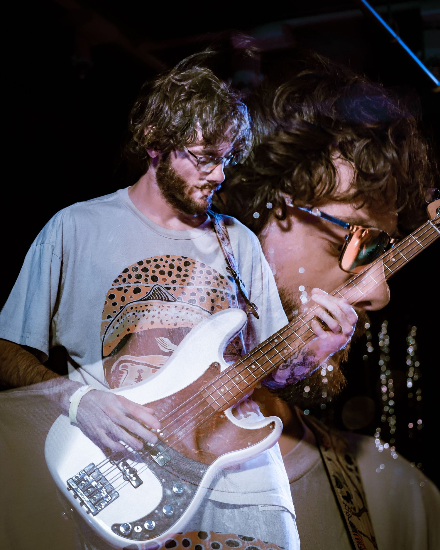 Two curly-haired musicians play bass guitar together under stage lights.