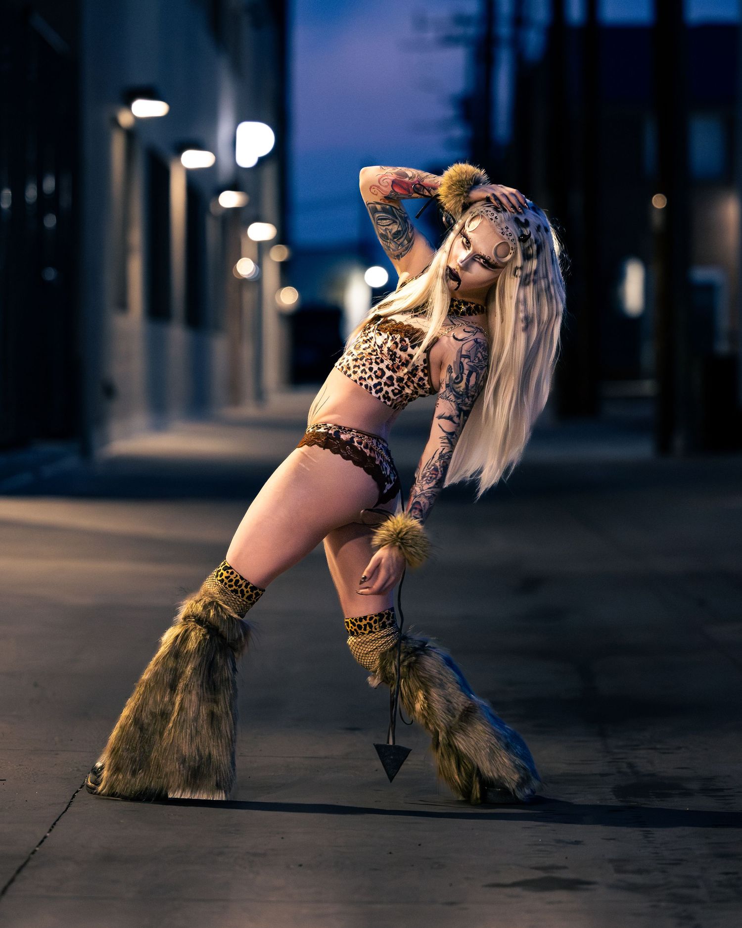 Tattooed blonde woman in leopard print outfit with fur leg warmers posing dramatically in night alley.