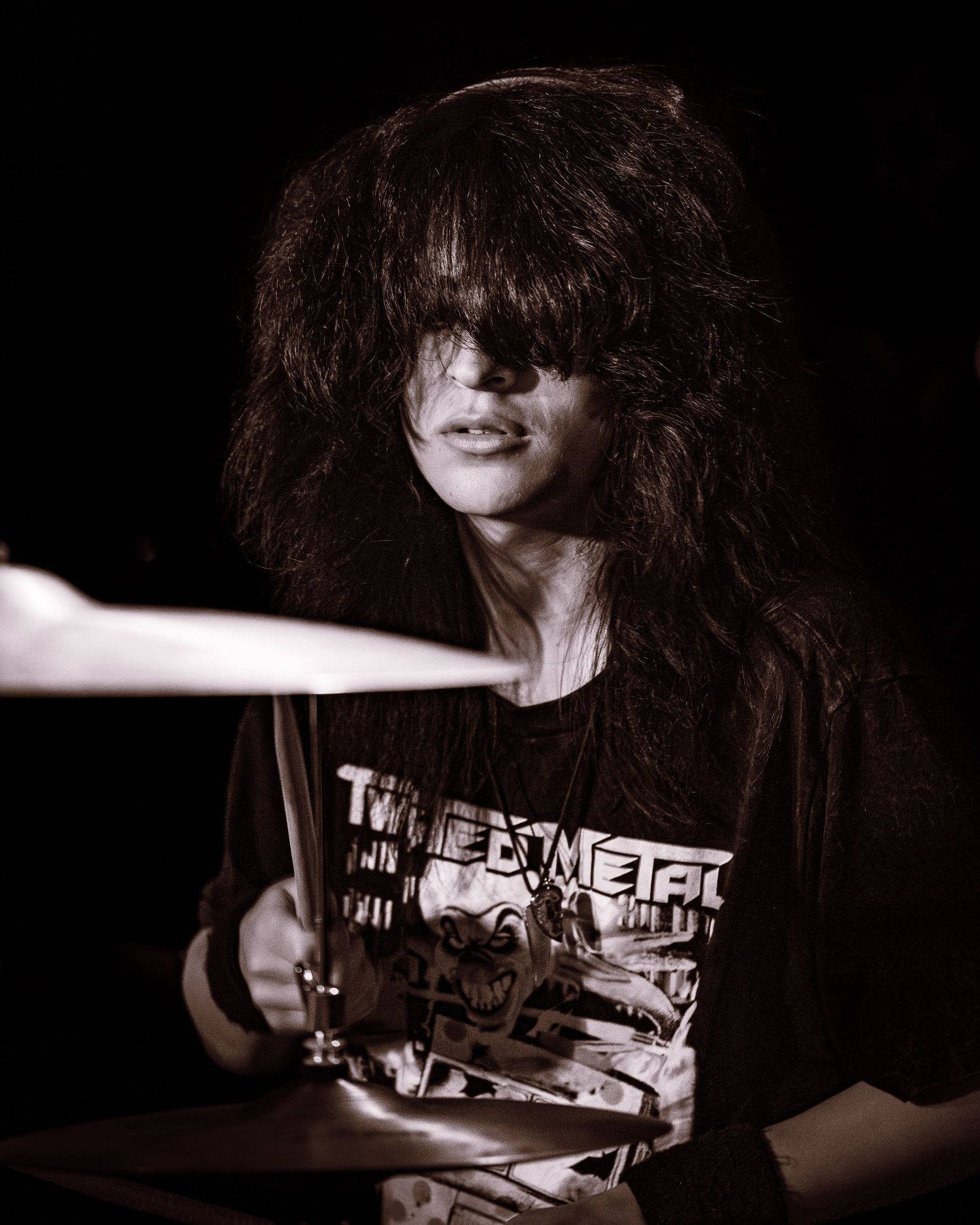 Long-haired drummer performing on stage in black metal band t-shirt in black and white photo.