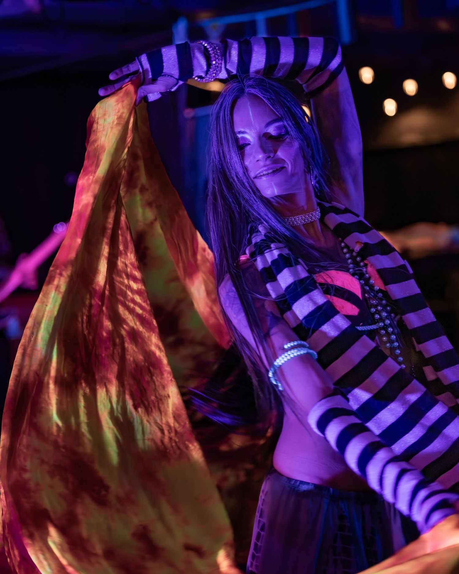Woman belly dancing under colorful neon lights wearing striped top and flowing tie-dye veil.