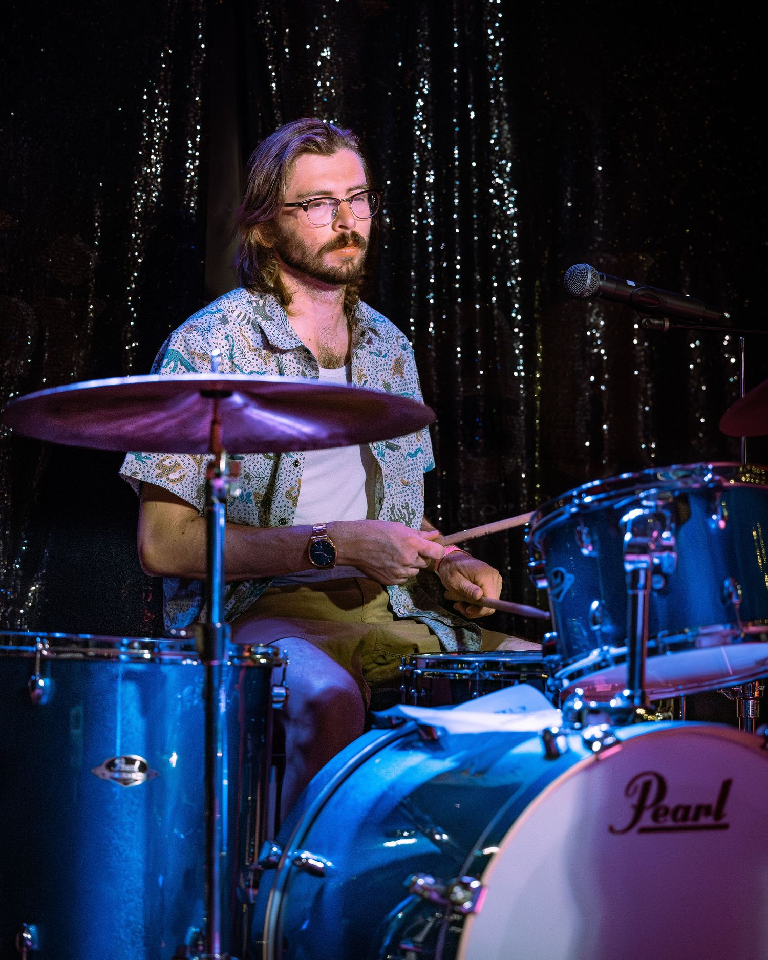 A bearded musician with glasses playing Pearl drums on a dimly lit stage.