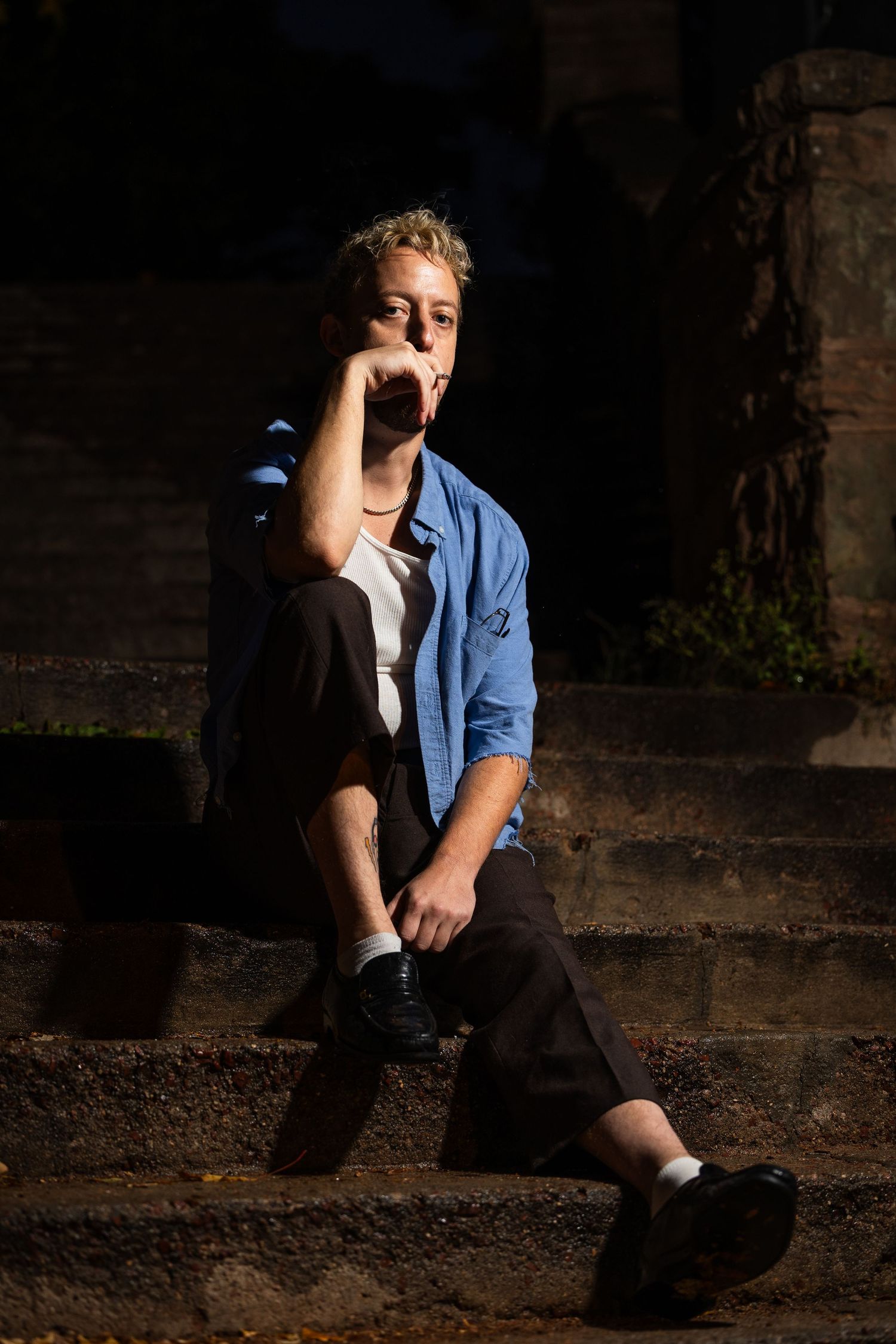A person in a blue shirt and black pants sits on stone steps in dramatic lighting with shadows.