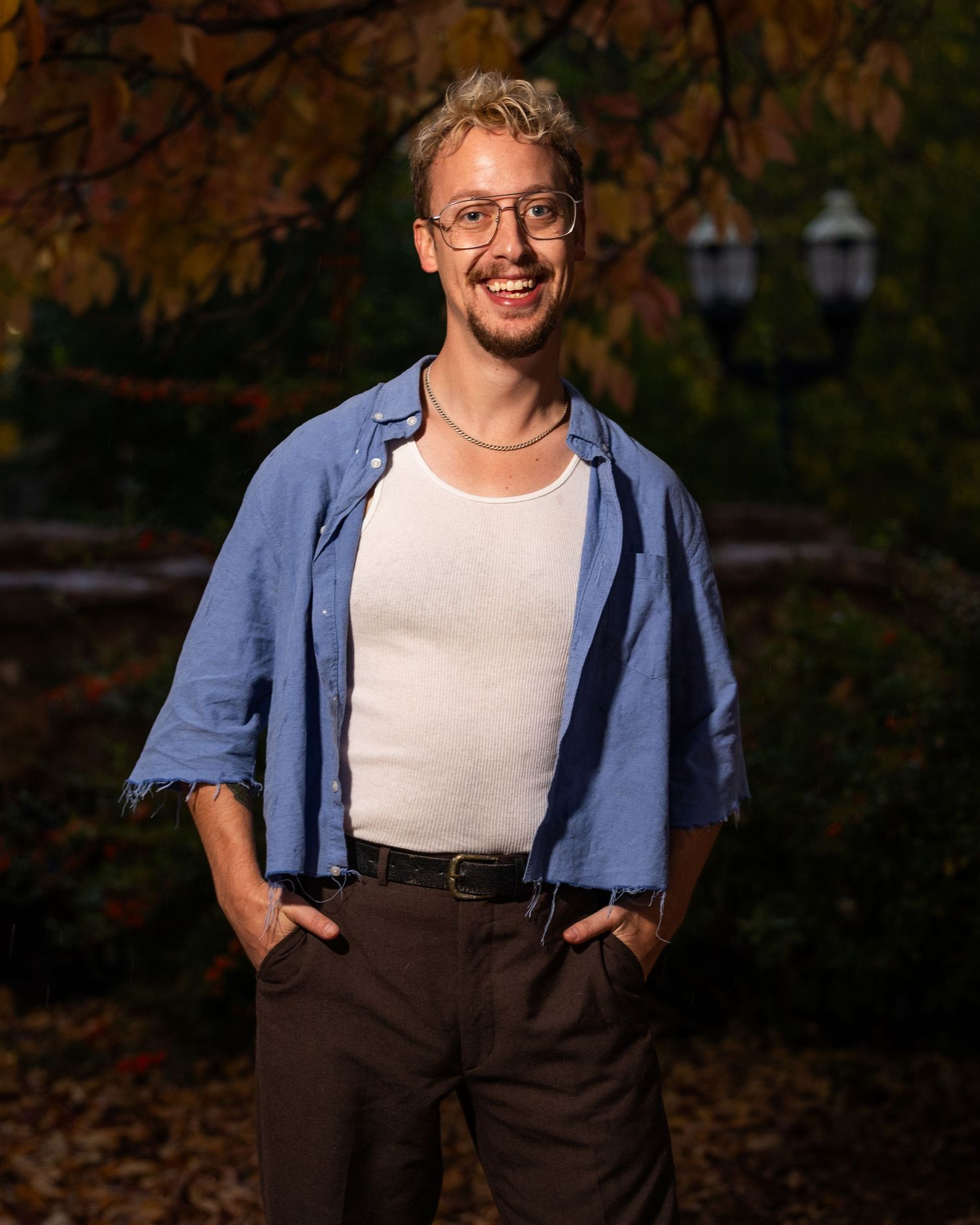 Person in blue denim shirt and dark suspenders poses outdoors during golden hour lighting.