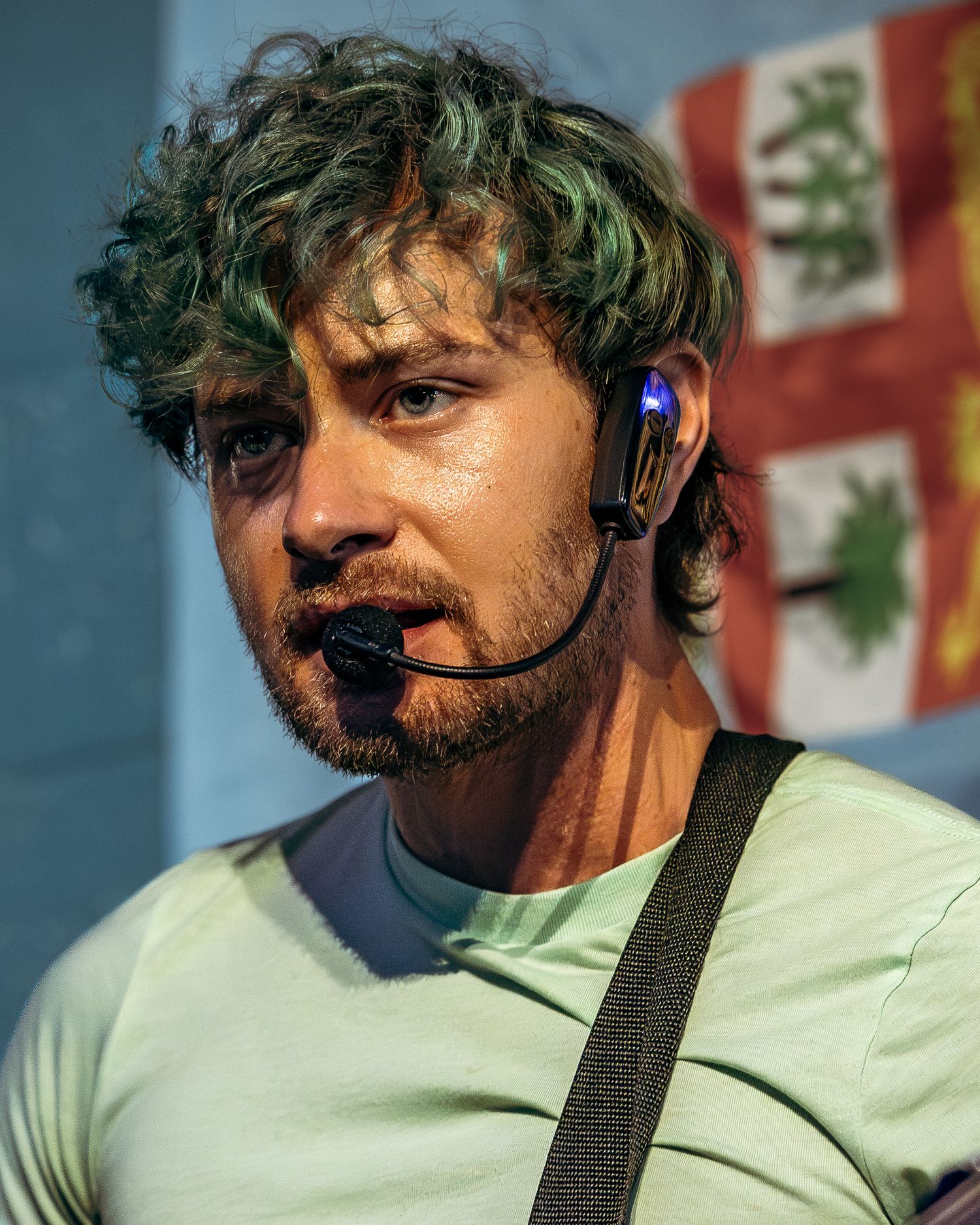 A performer wearing a white t-shirt and headset microphone stands against a colorful background during a show.