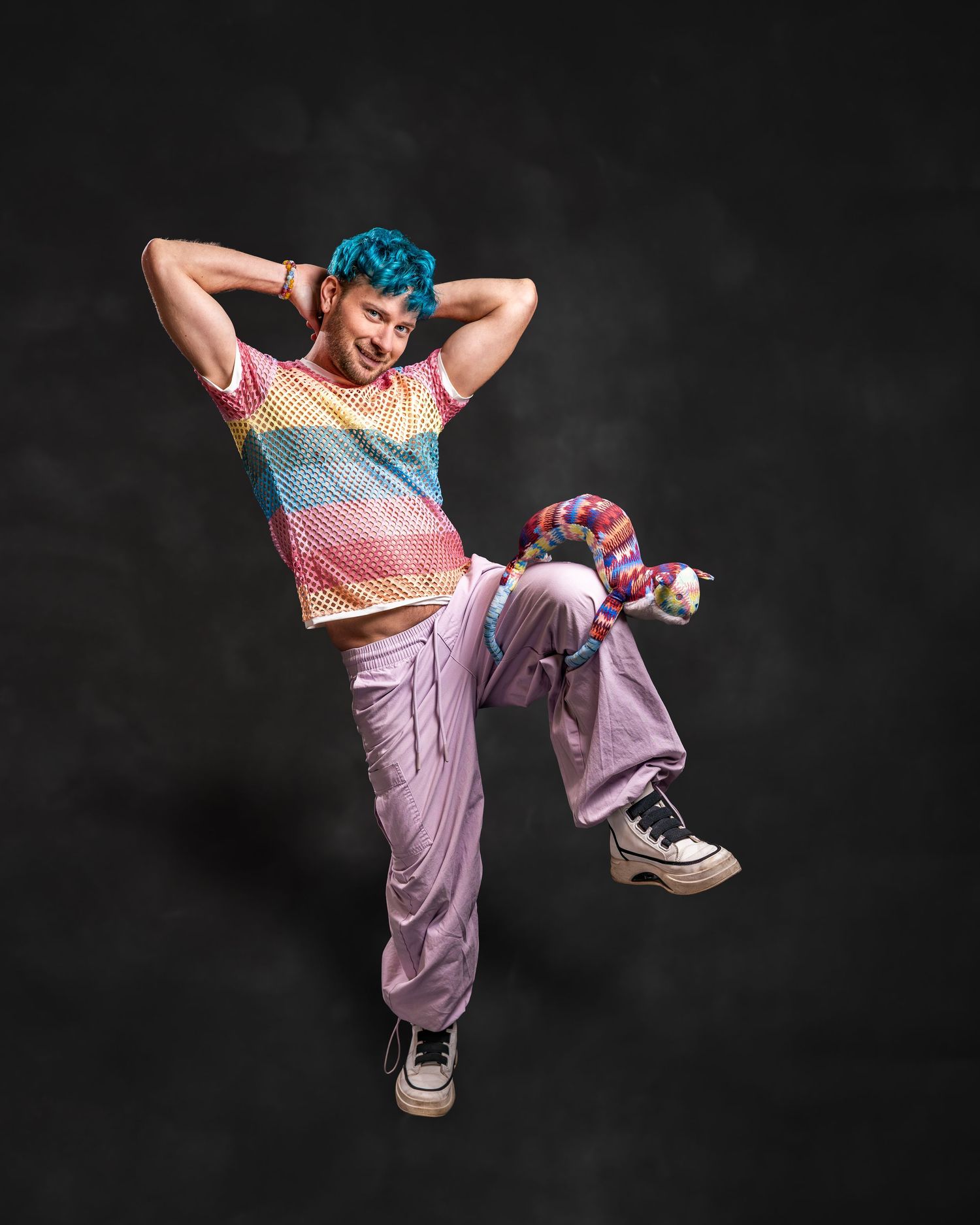 Person with blue hair dancing in rainbow mesh top and lavender pants against dark background.