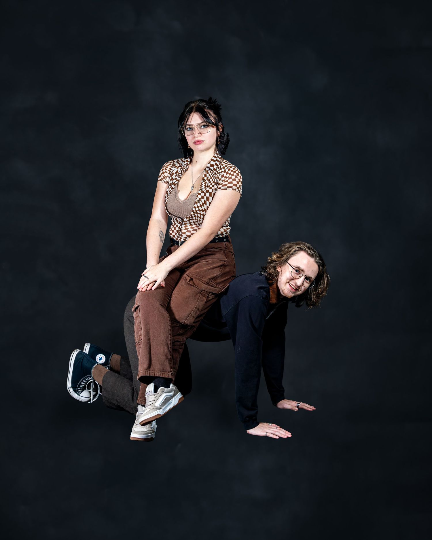 Two young people pose dramatically in a studio, one standing over the other who is on all fours.