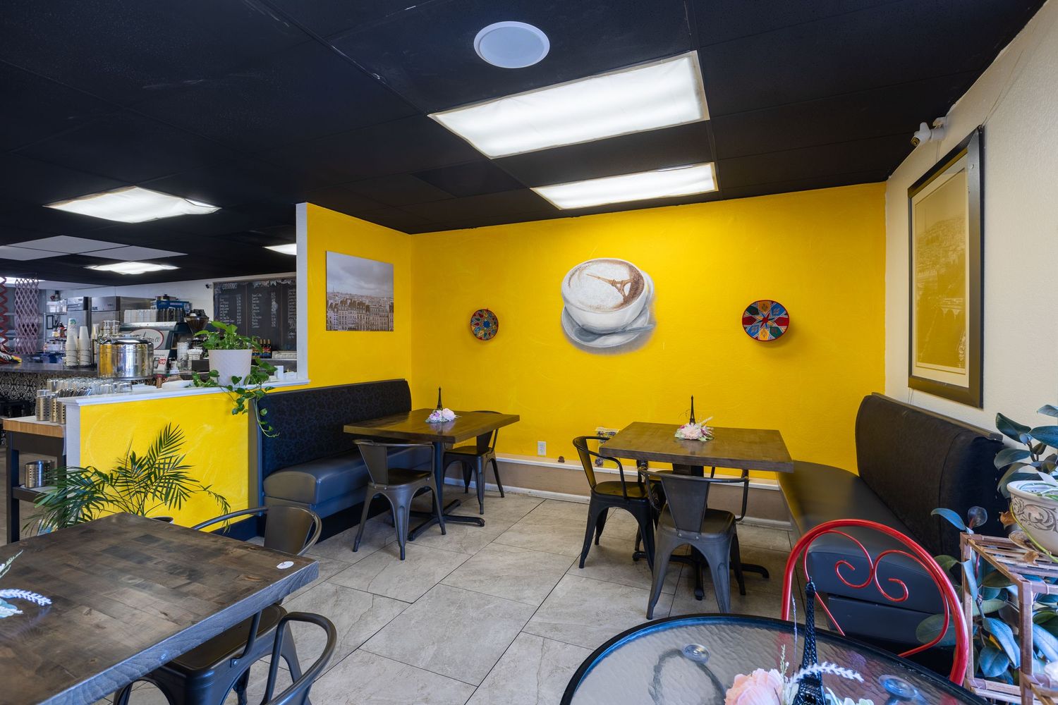 Cozy cafe dining area with bold yellow walls, black metal tables and chairs, artwork, and tile flooring.