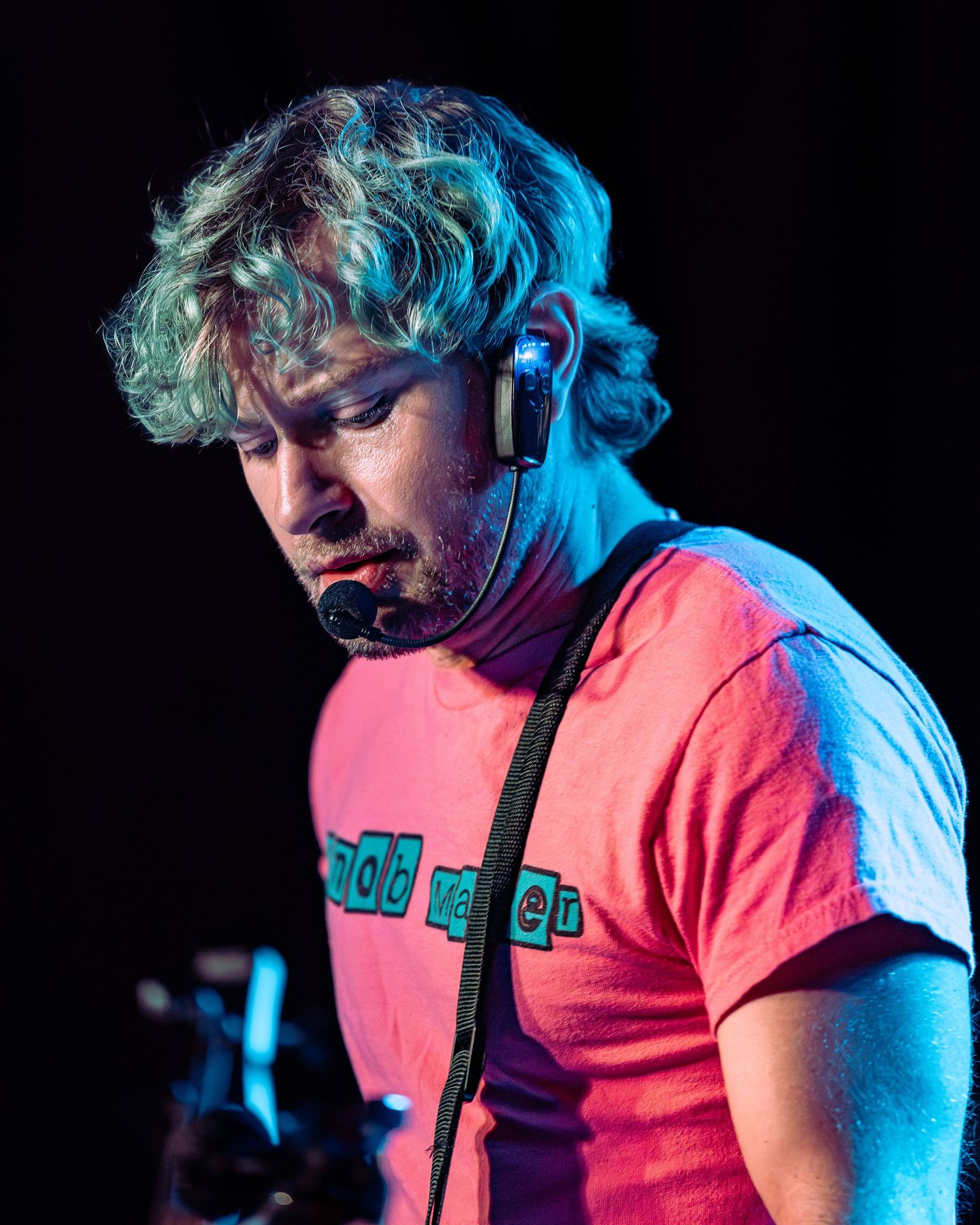 Curly-haired guitarist in red t-shirt focuses downward while playing on a dark concert stage.
