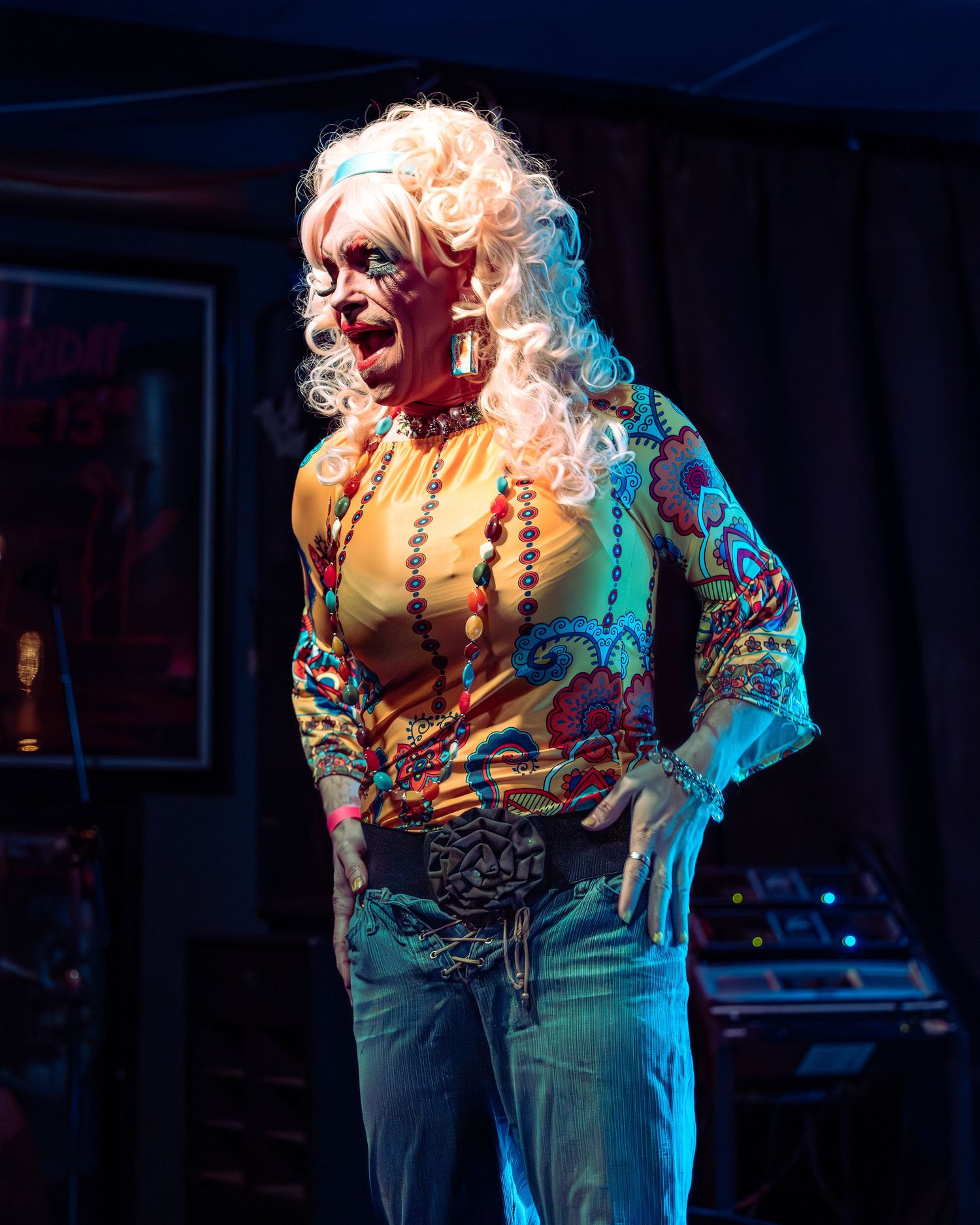 Performer in blonde curly wig and colorful floral top singing on stage under warm lighting.