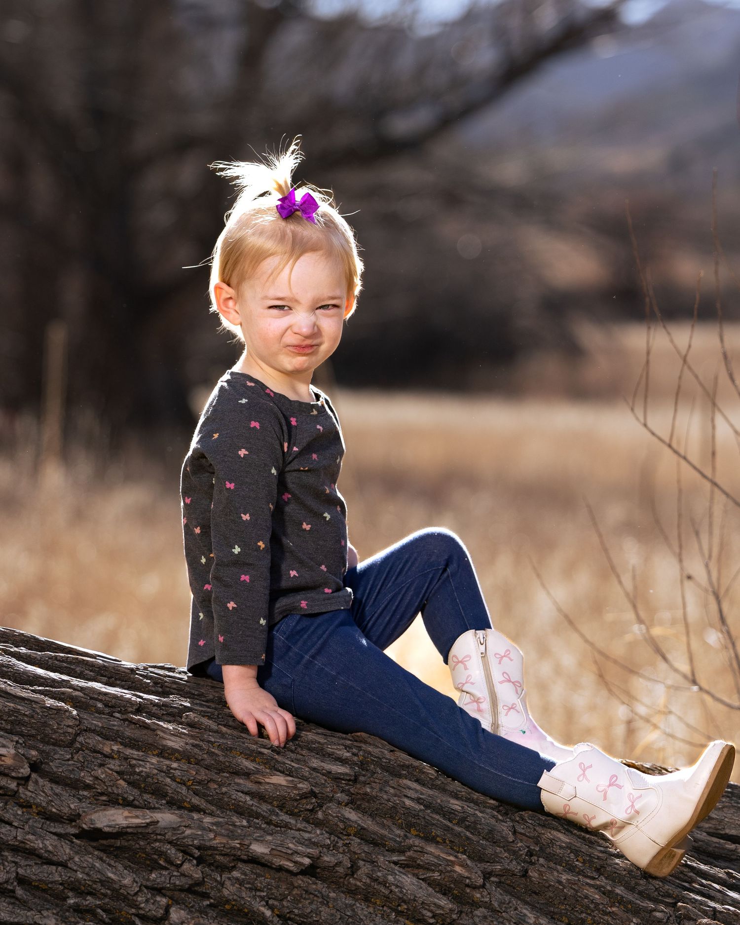 Young blonde girl in dark floral top and jeans sits on a log outdoors, wearing pink boots and a purple bow.