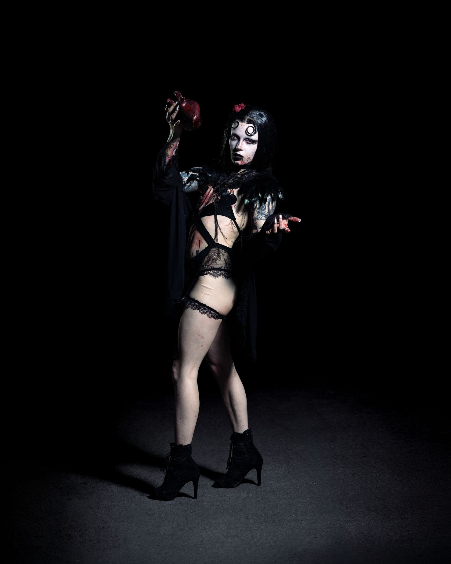 Person in dark gothic Halloween costume with dramatic makeup, black cape, lace lingerie, and heels poses dramatically.