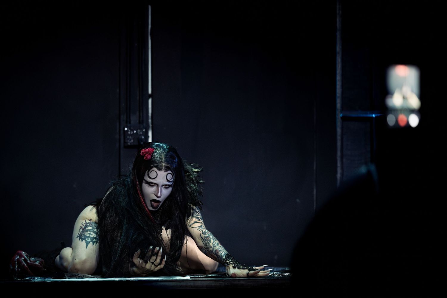 Performer with gothic makeup and tattoos leans over a surface on a dark dramatic stage.
