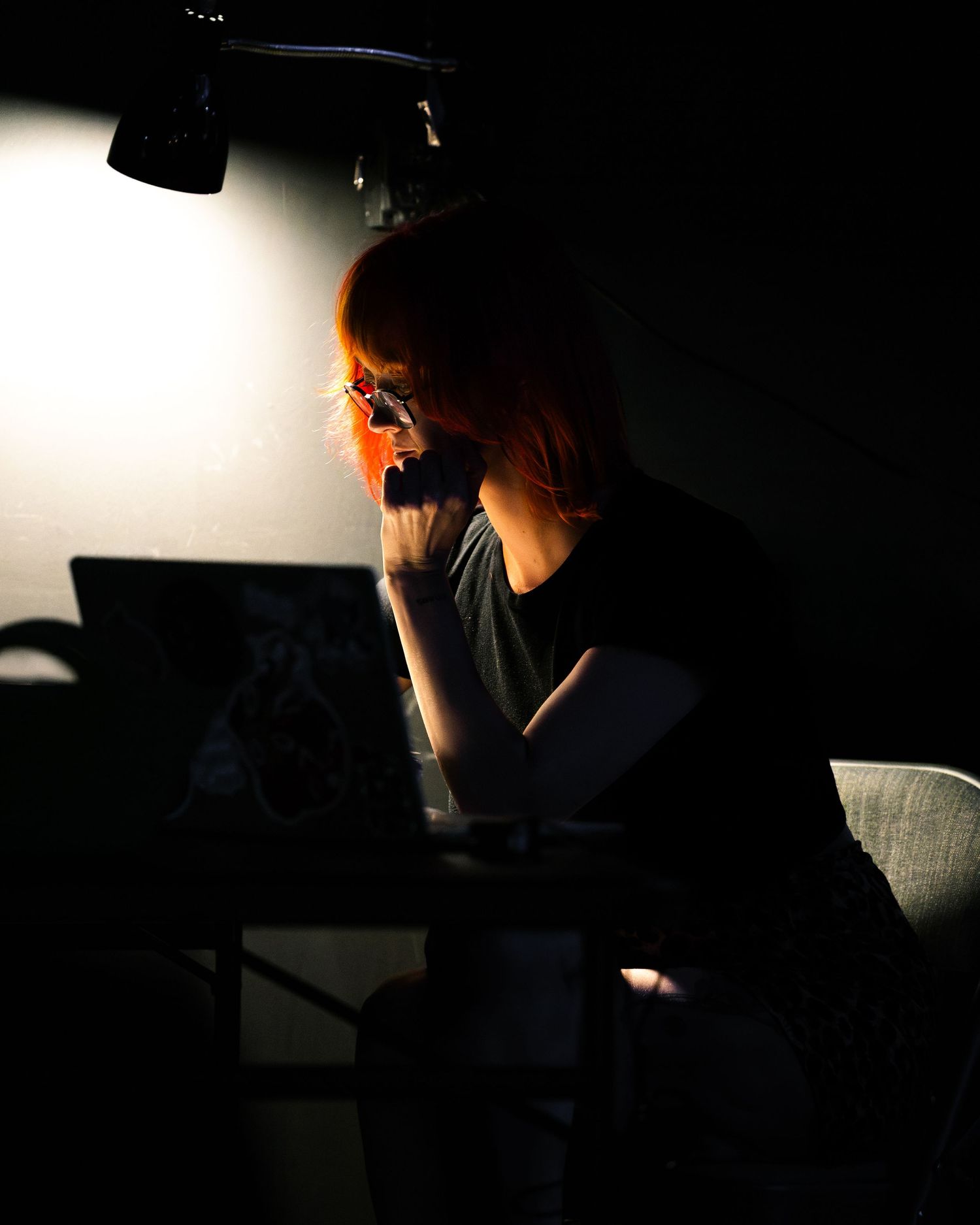 A red-haired woman sits under a spotlight in a dark room, thoughtfully resting her hand near her face.