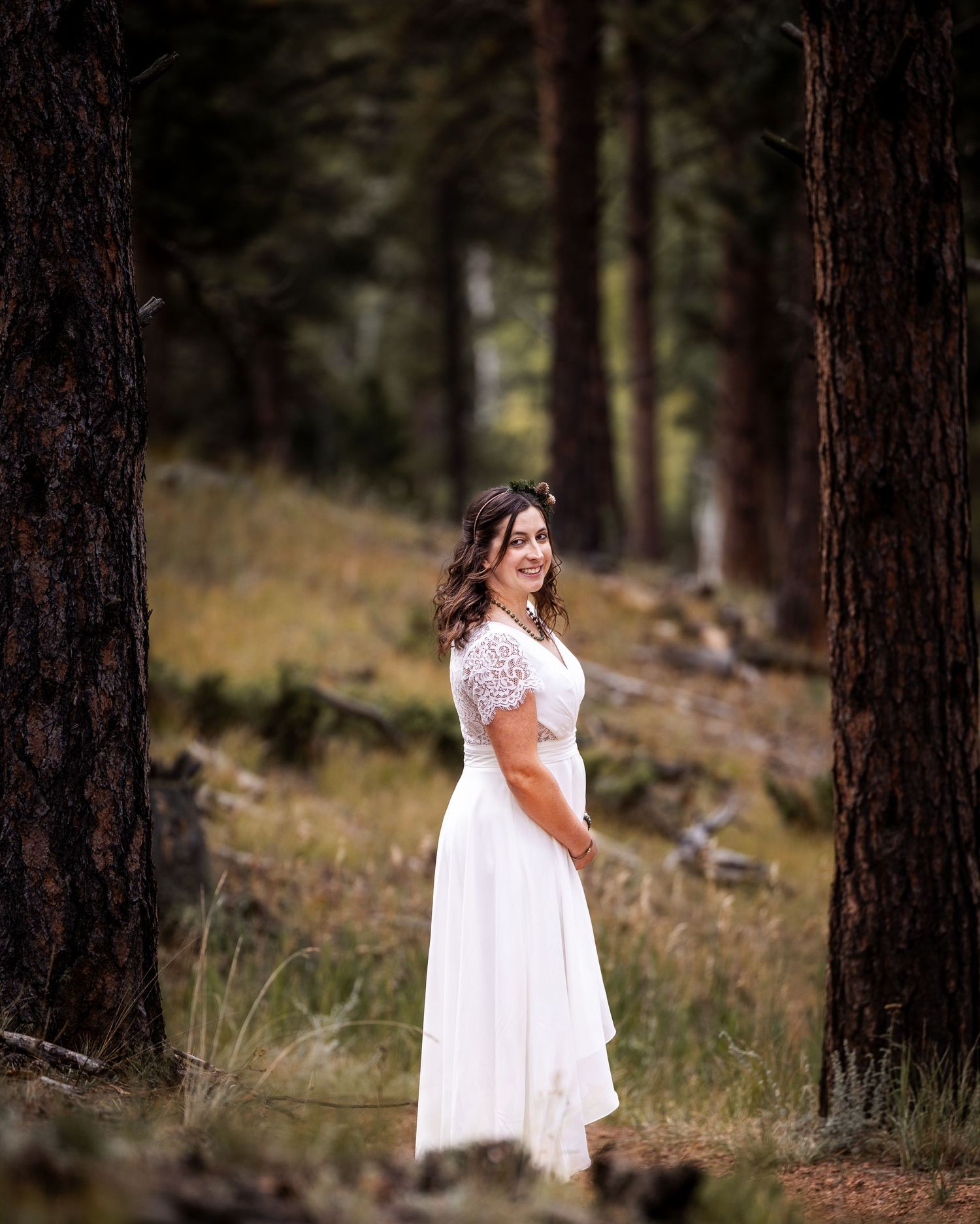 A white lace dress flows elegantly against a backdrop of tall pine trees in a forest setting.